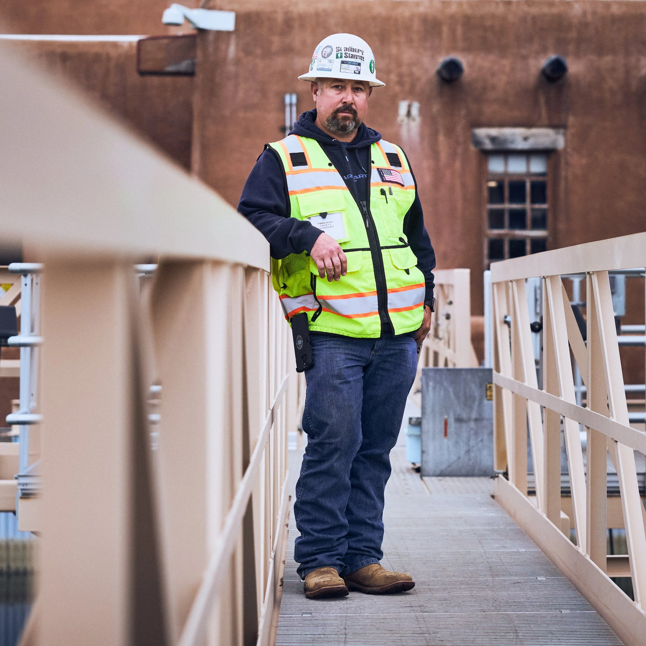 A construction worker in a white hard hat and high-visibility safety vest standing on a wooden bridge or platform, with a brick building in the background.