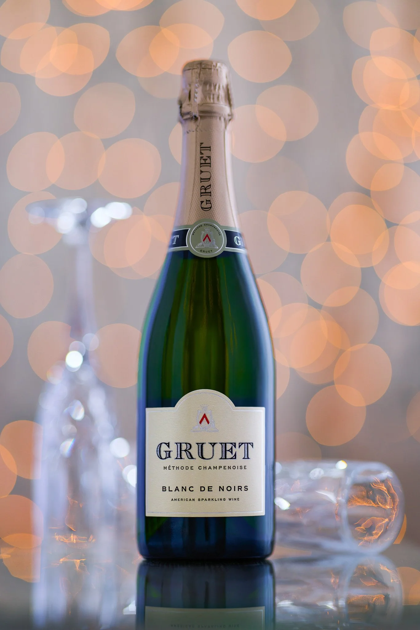 A bottle of Gruet Blanc de Noirs sparkling wine with a blurred background of warm-toned bokeh lights and a partially blurred wine glass to the left.