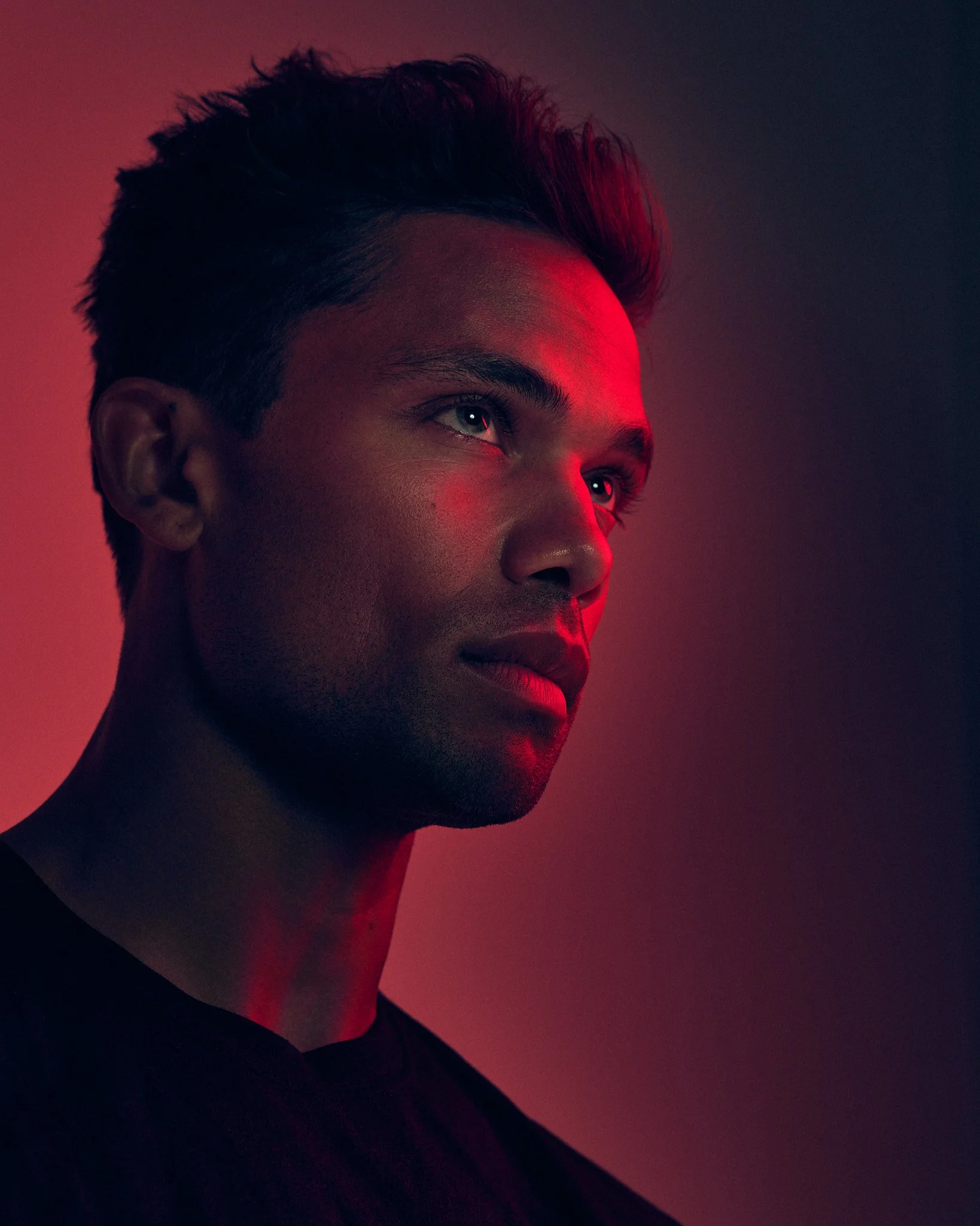 Close-up of a young man's face with dramatic red lighting on the right side, dark background, short styled hair, and a calm expression.