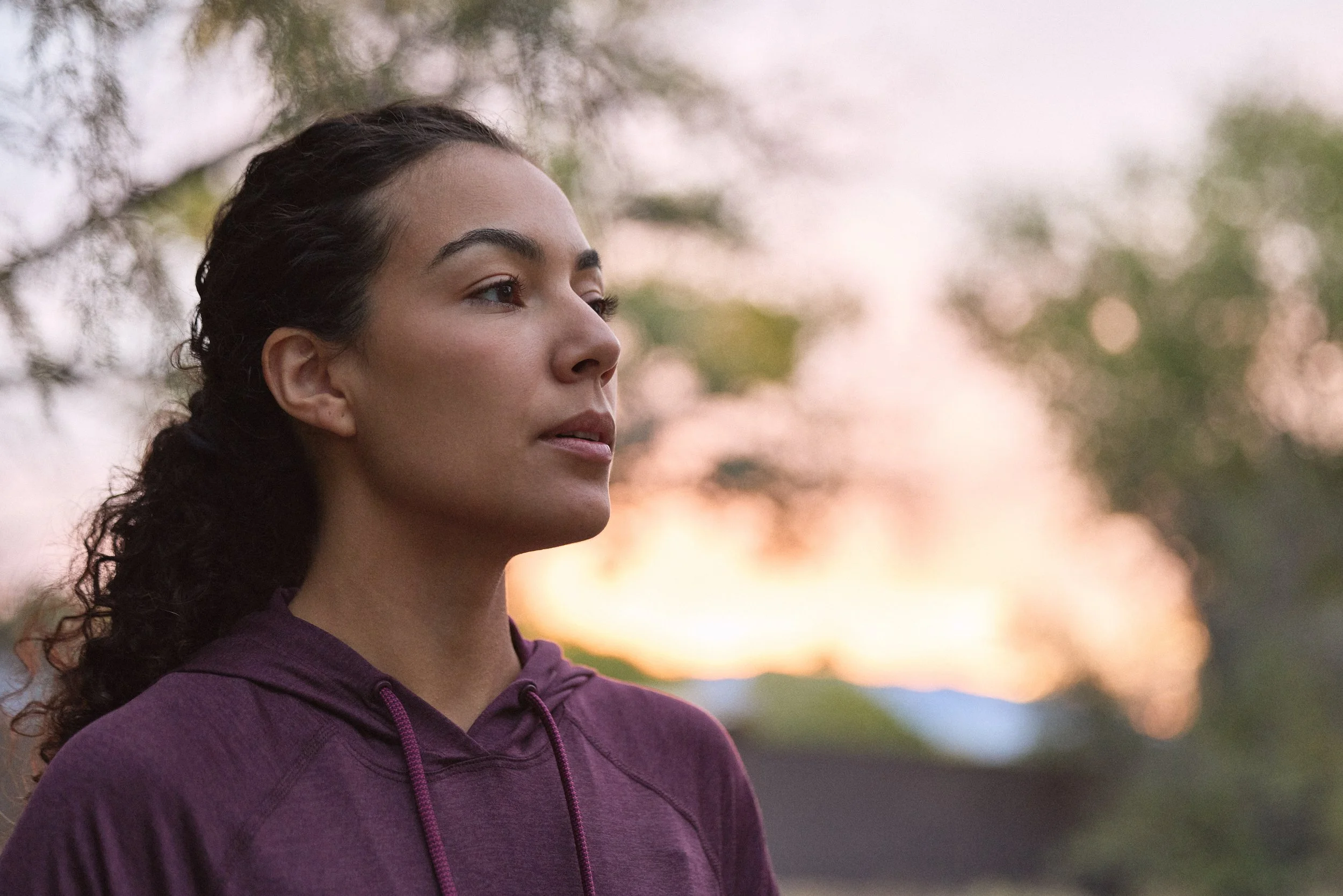 A young woman with dark, curly hair tied back, wearing a purple hoodie, appears to be outdoors during sunset or sunrise, with blurred trees and a colorful sky in the background.