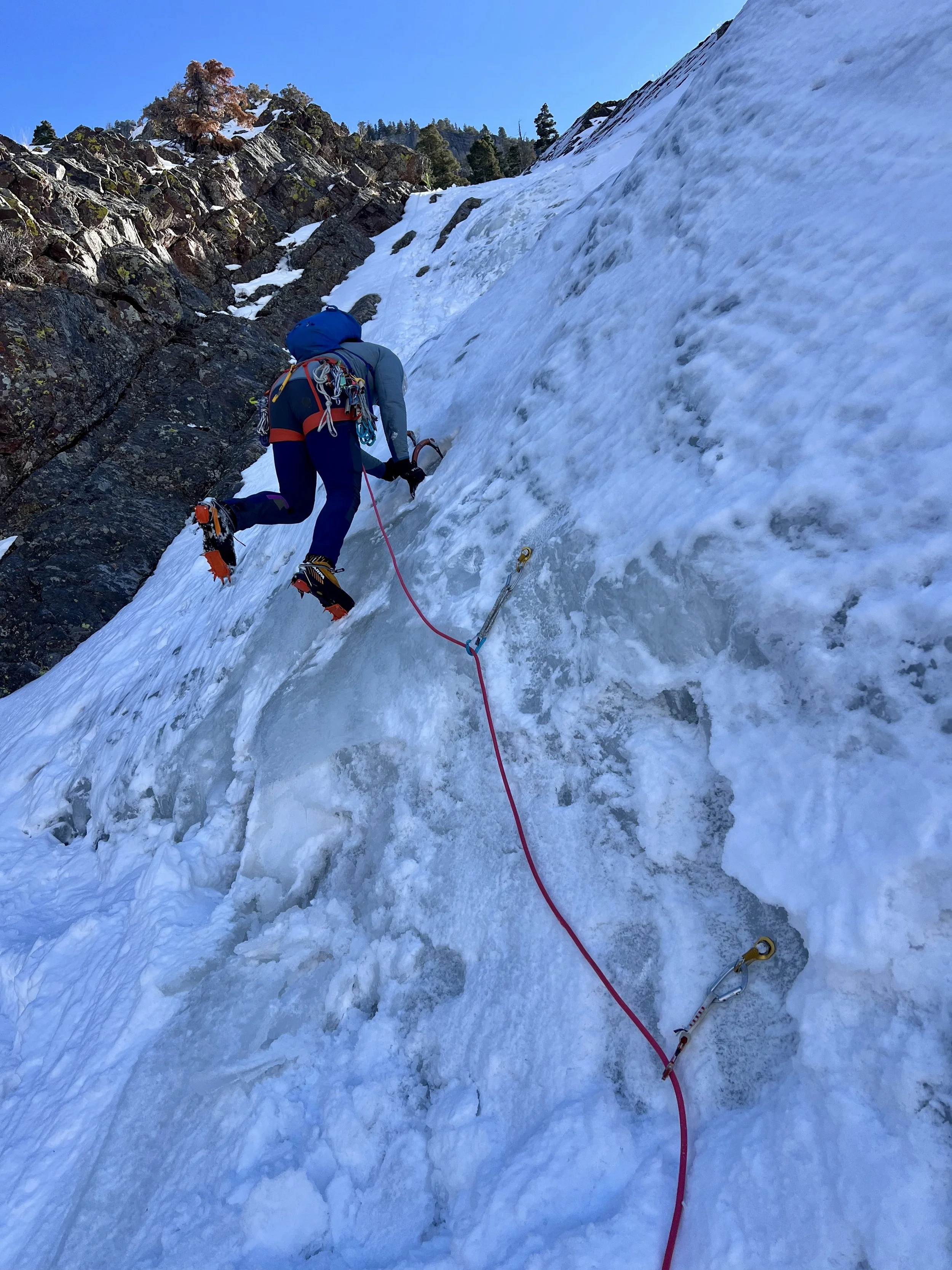 Person wearing winter gear ice climbing on a frozen waterfall or ice-covered rock face with a climbing harness and safety ropes.