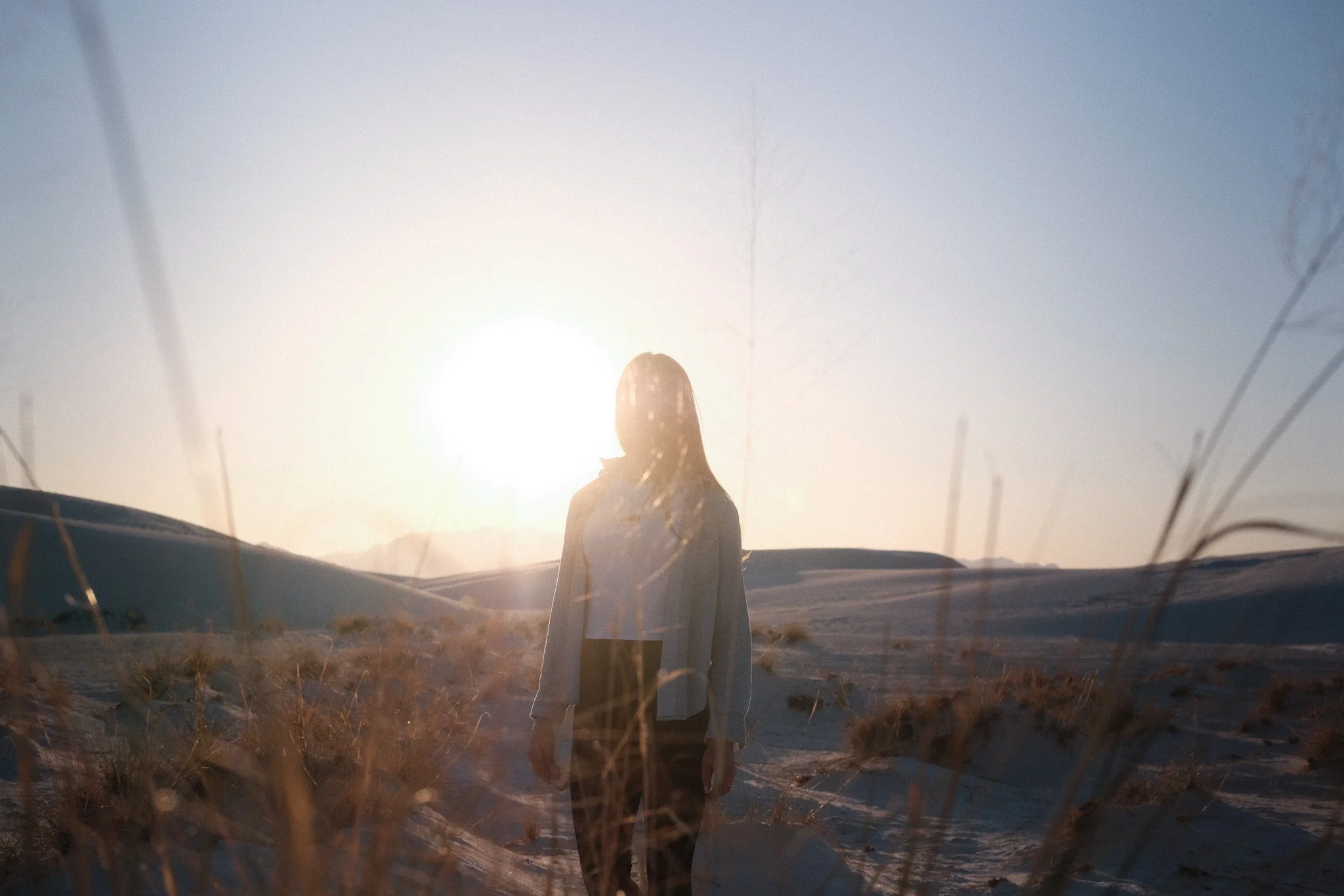 Person standing outdoors in a field at sunset, with a sunlit sky and mountains in the background.