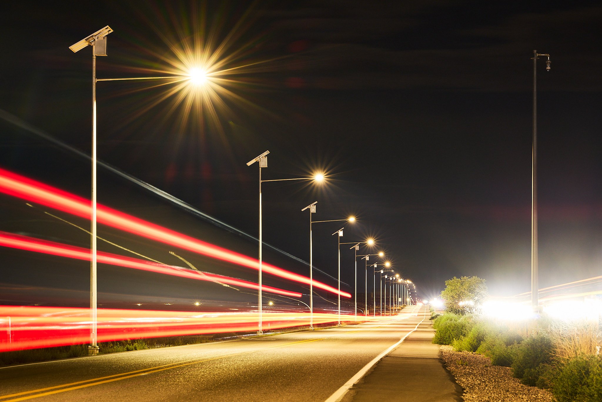 Nighttime scene of a road with multiple evenly spaced streetlights, some equipped with solar panels, and streaks of red and white light from moving vehicles.