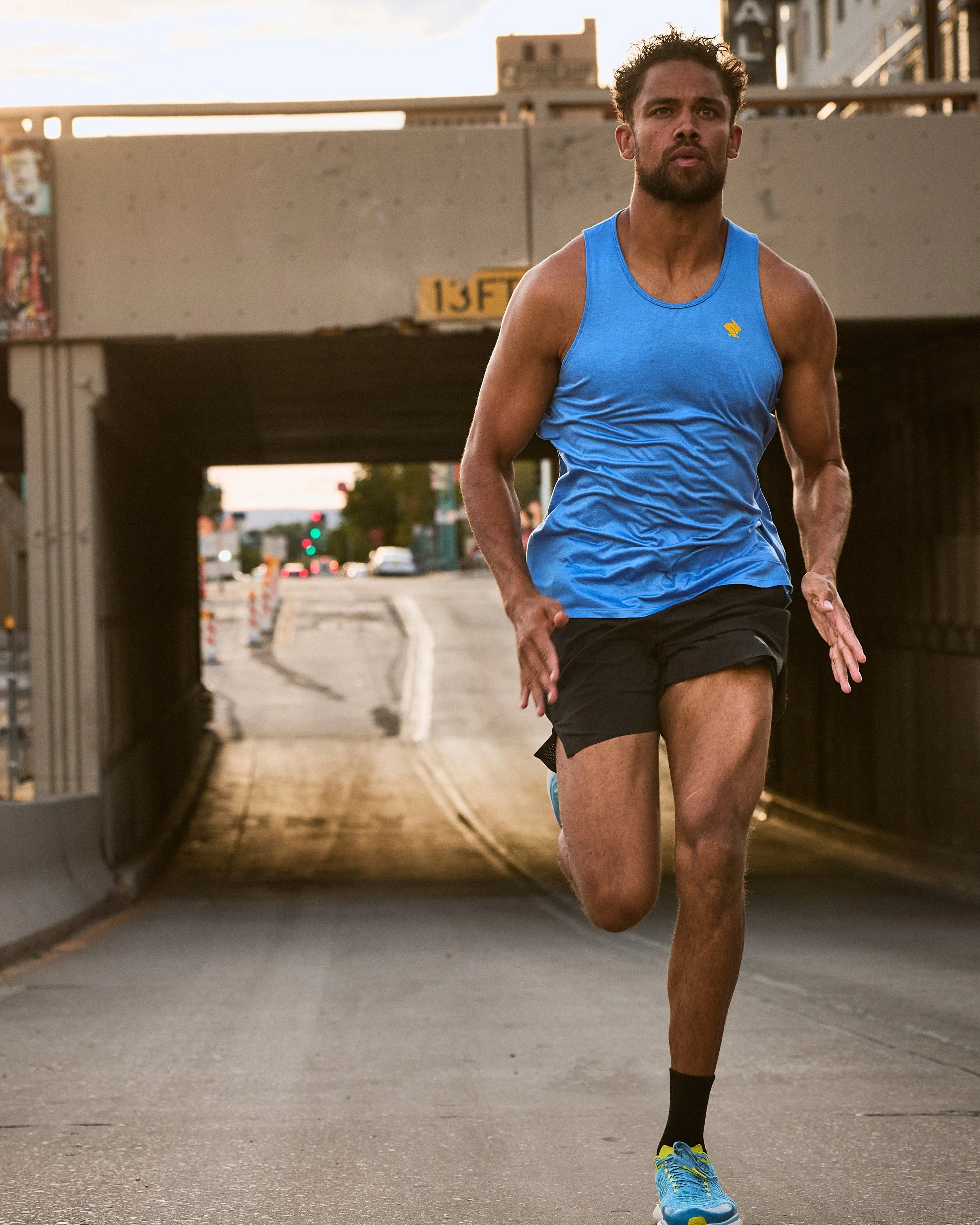 A man running on a city street under a bridge at sunset. He is wearing a blue sleeveless shirt, black shorts, and blue running shoes with yellow accents.