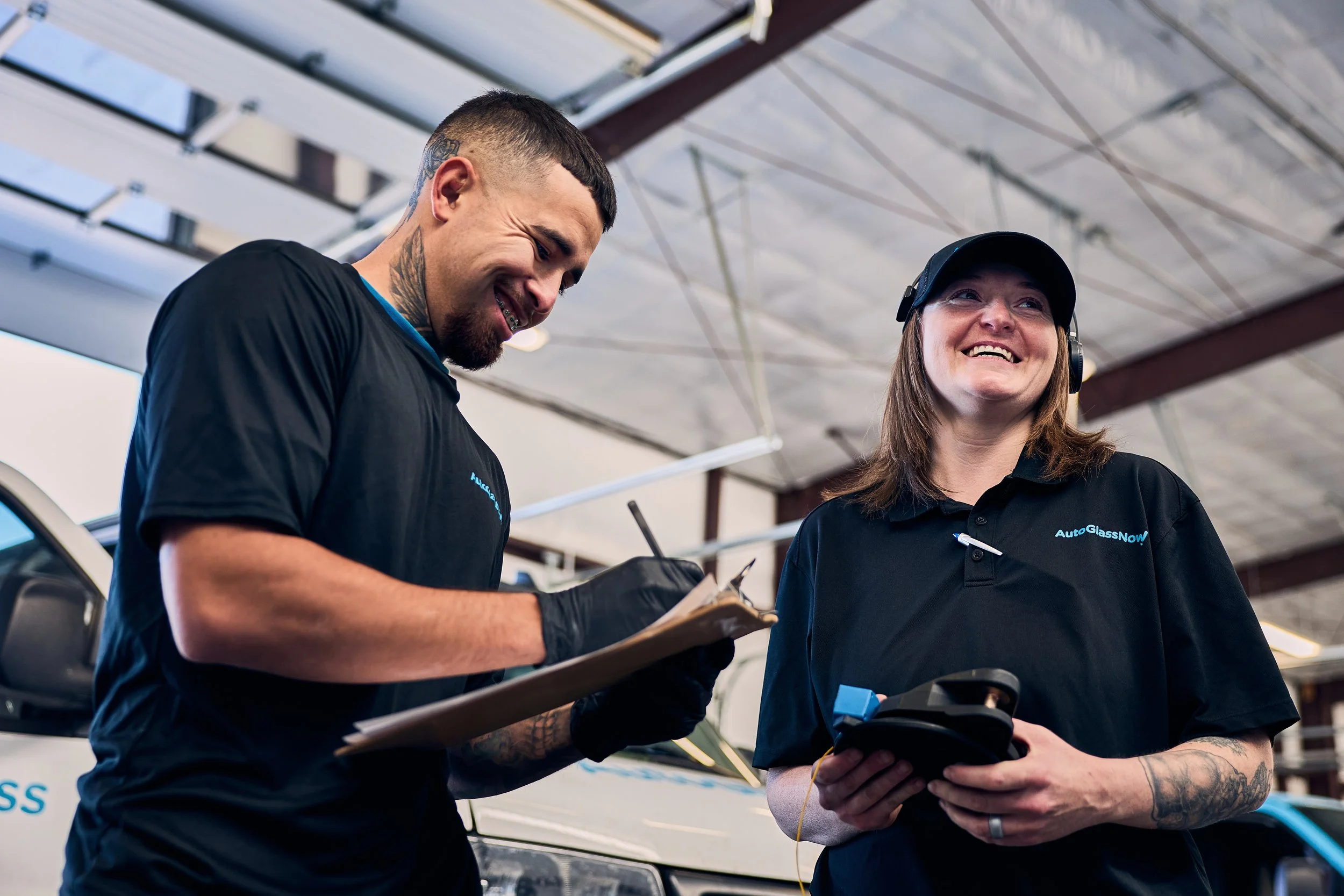 Two technicians working together at an auto glass repair shop, both wearing black shirts with 'Auto Glass Now' logos, discussing work details and smiling.