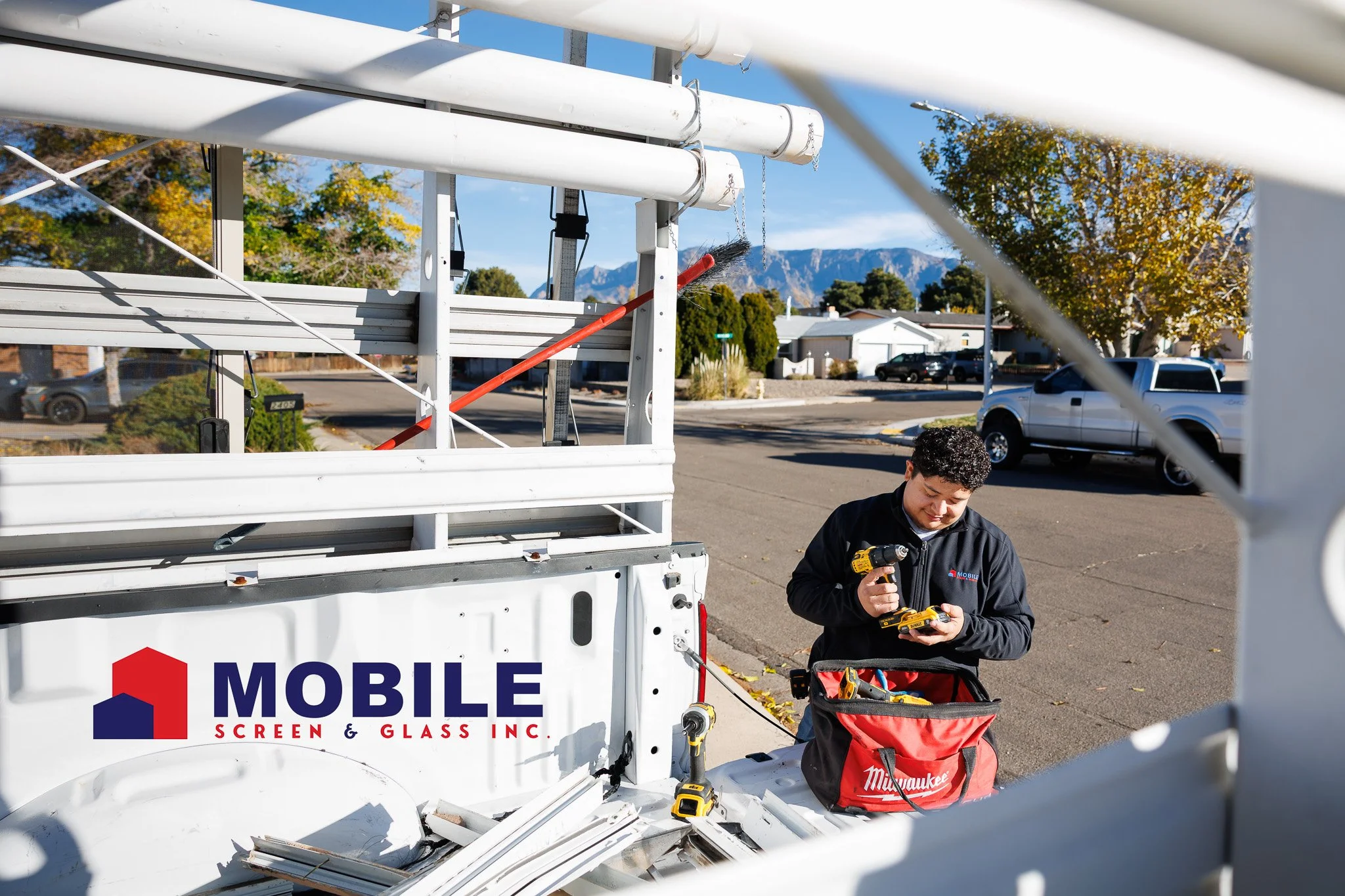 A technician working on a mobile screen and glass repair truck, wearing a black jacket and using a yellow cordless drill, with trees and mountains in the background on a clear day.