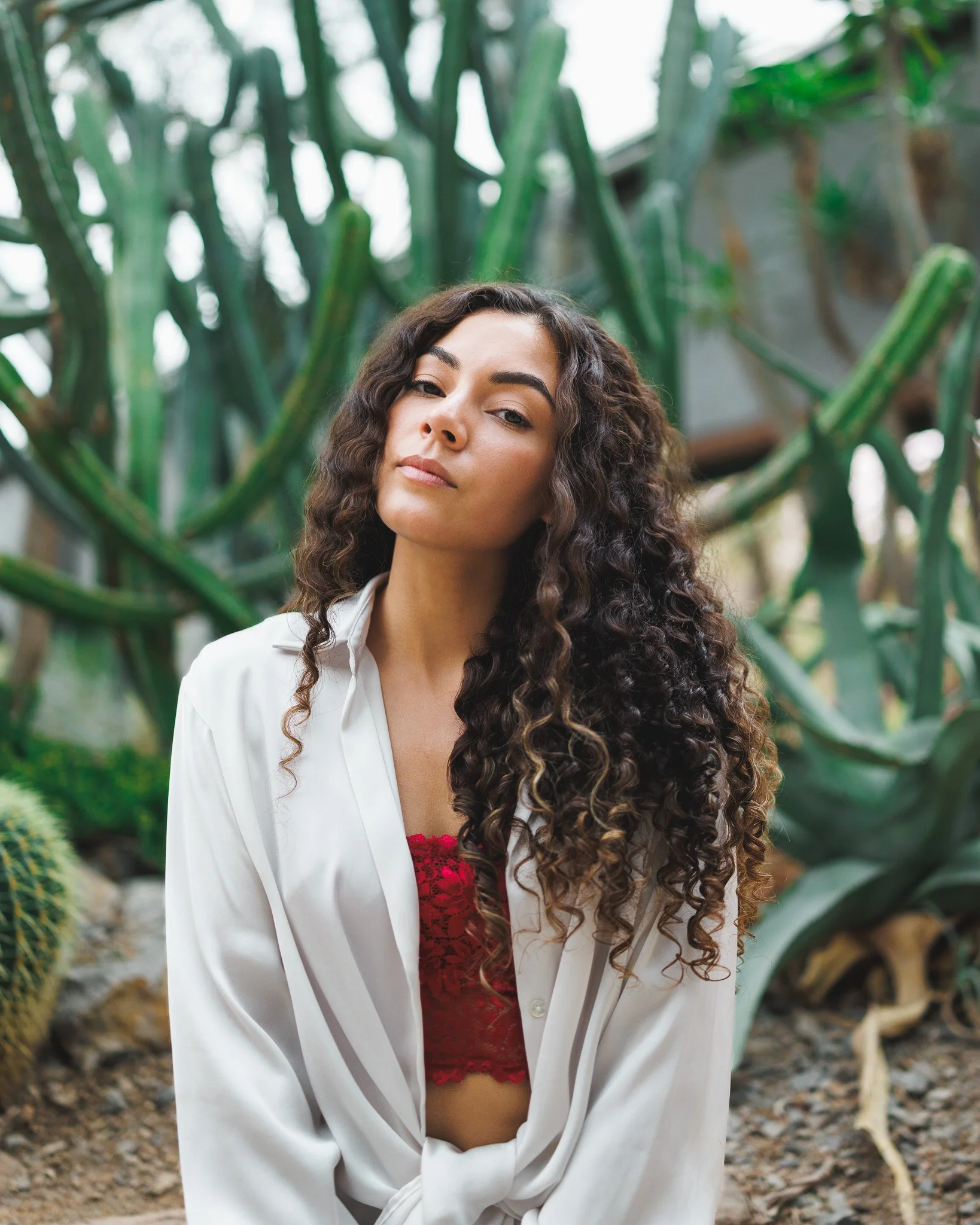 A young woman with curly brown hair and light skin posing outdoors among lush green plants, wearing a white blazer over a red lace top with a confident expression.