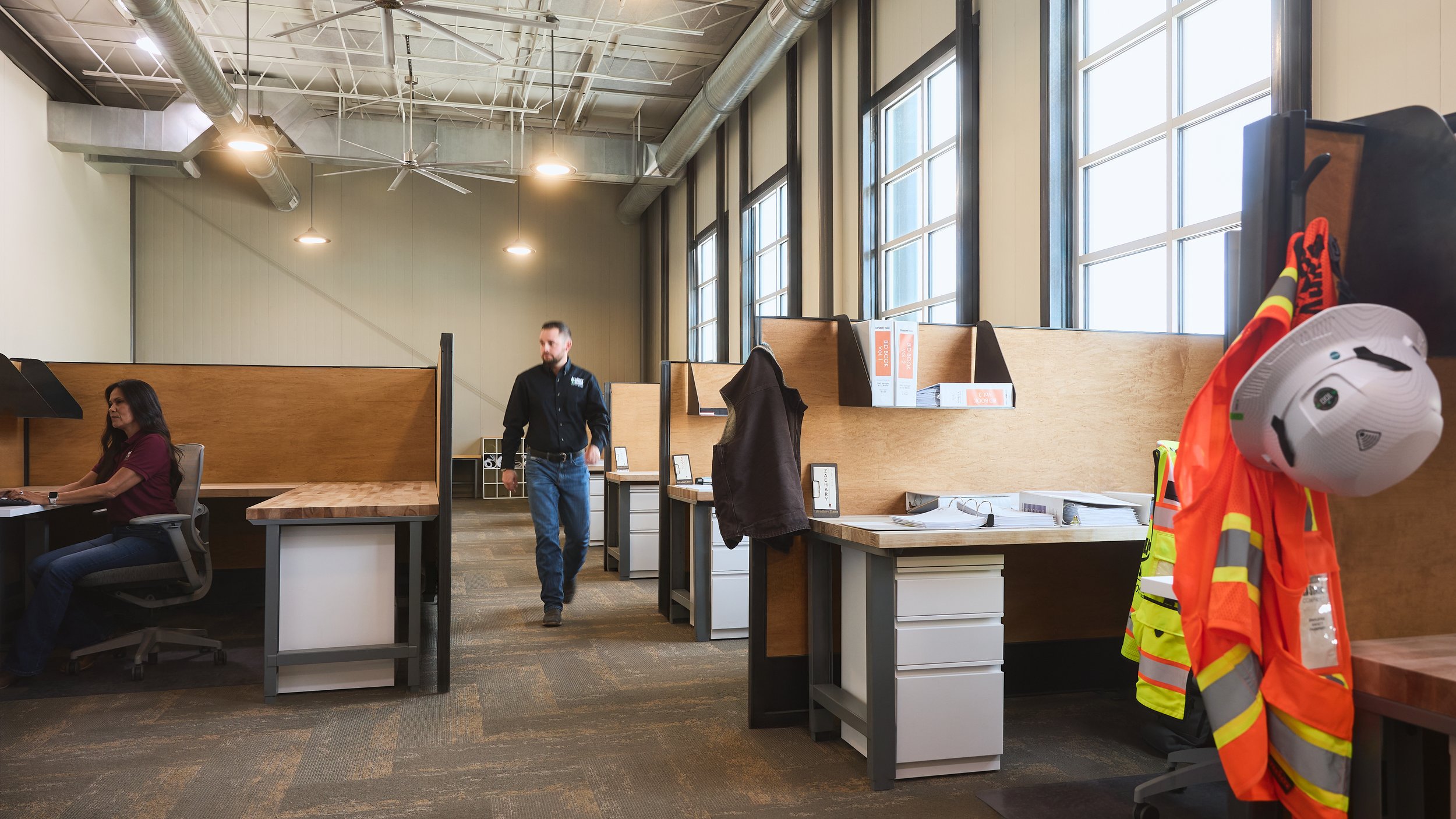 Office workspace with cubicles, a woman sitting at her desk, a man walking between desks, and safety gear hanging on a coat rack, including a white helmet and an orange safety vest.