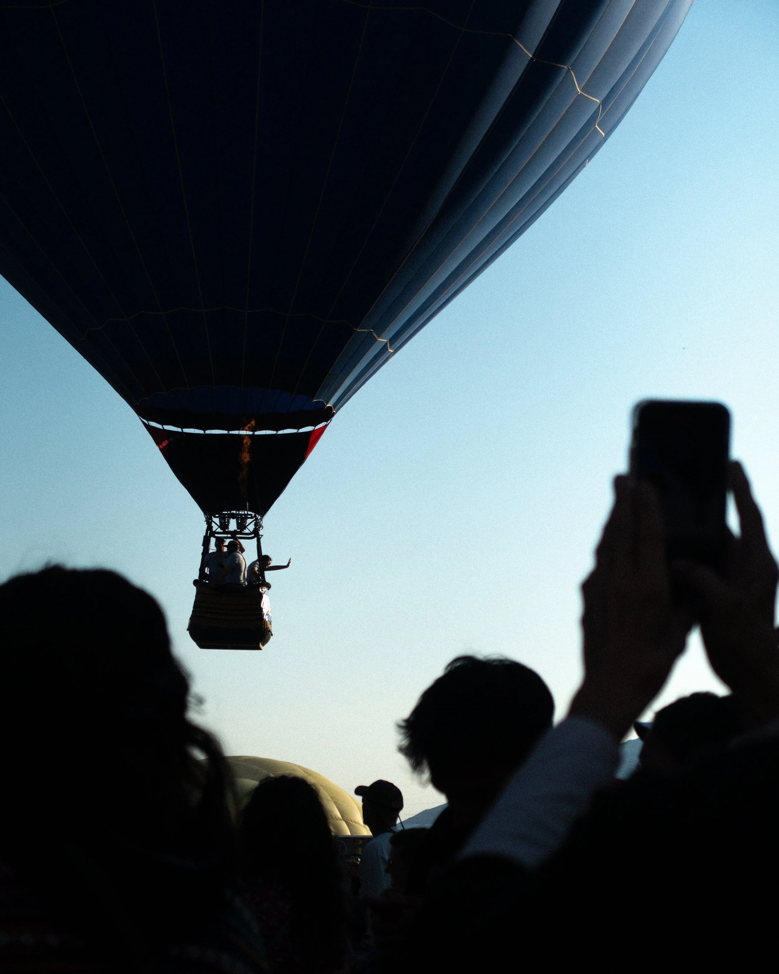 Hot air balloon flying with a crowd of people on the ground watching and taking photos.