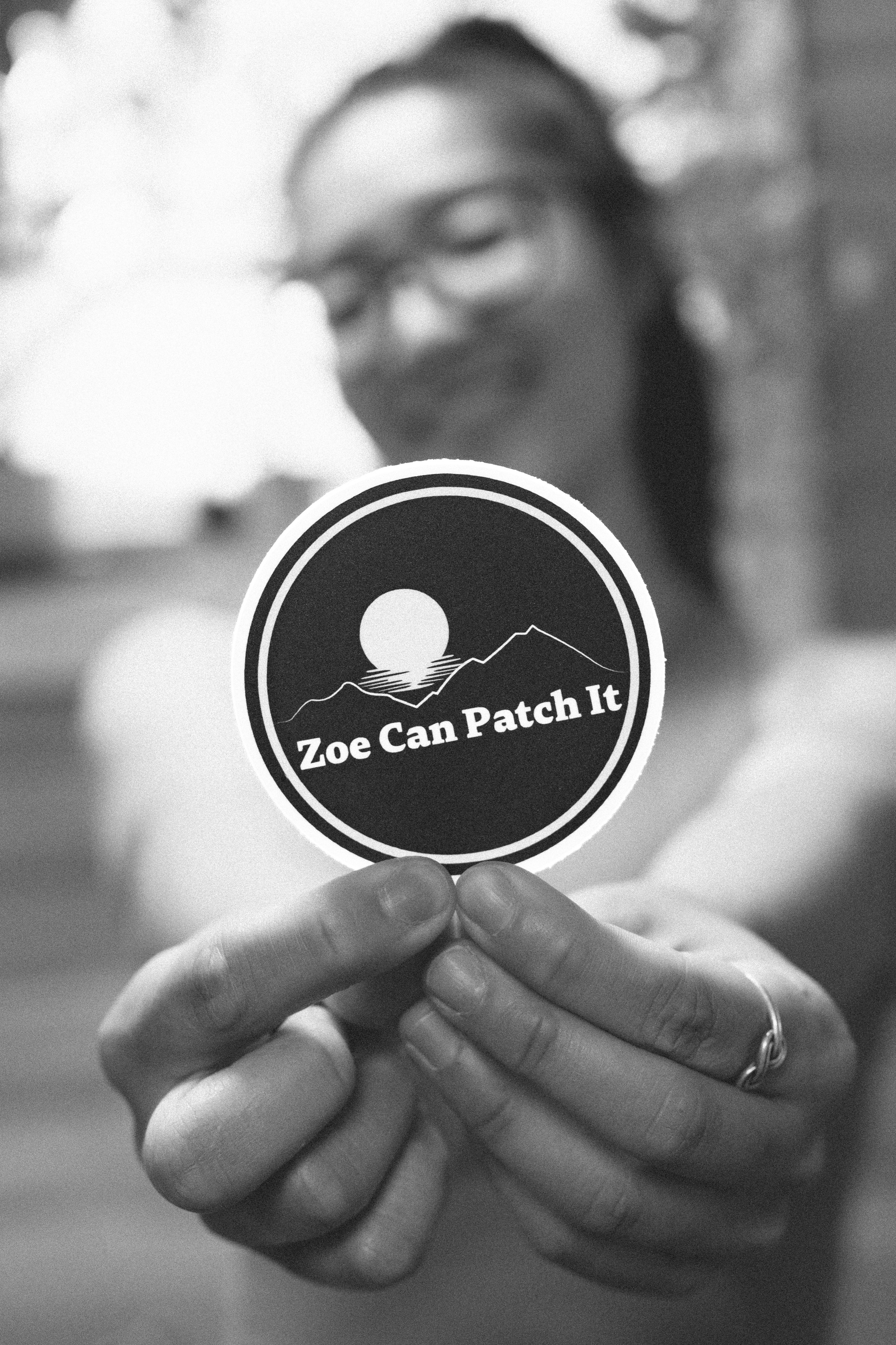 Person holding a round patch with a mountain and sun design, and the text 'Zoe Can Patch It' in black and white.
