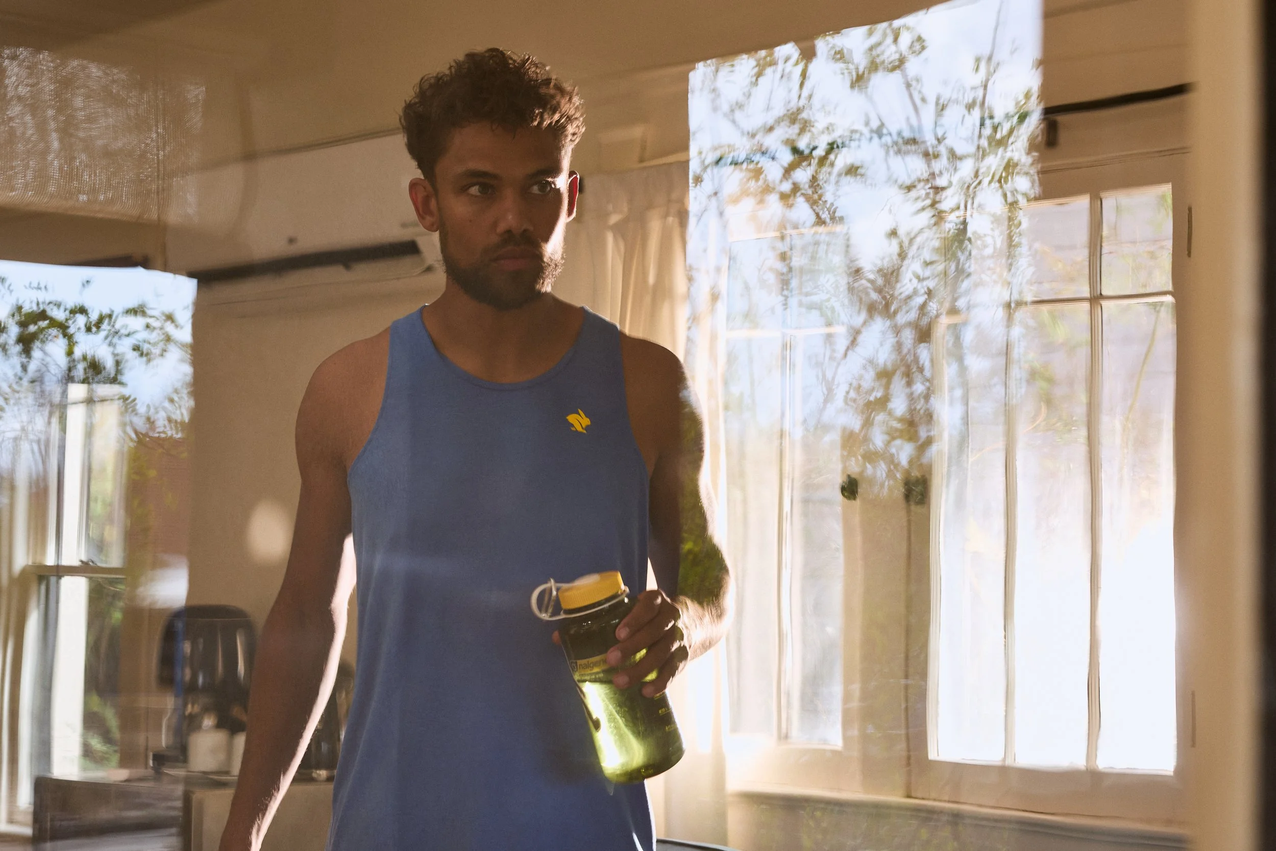 A young man with curly hair and a beard, wearing a blue tank top, holding a yellow-lidded water bottle, standing inside a room with sunlight streaming through the windows.