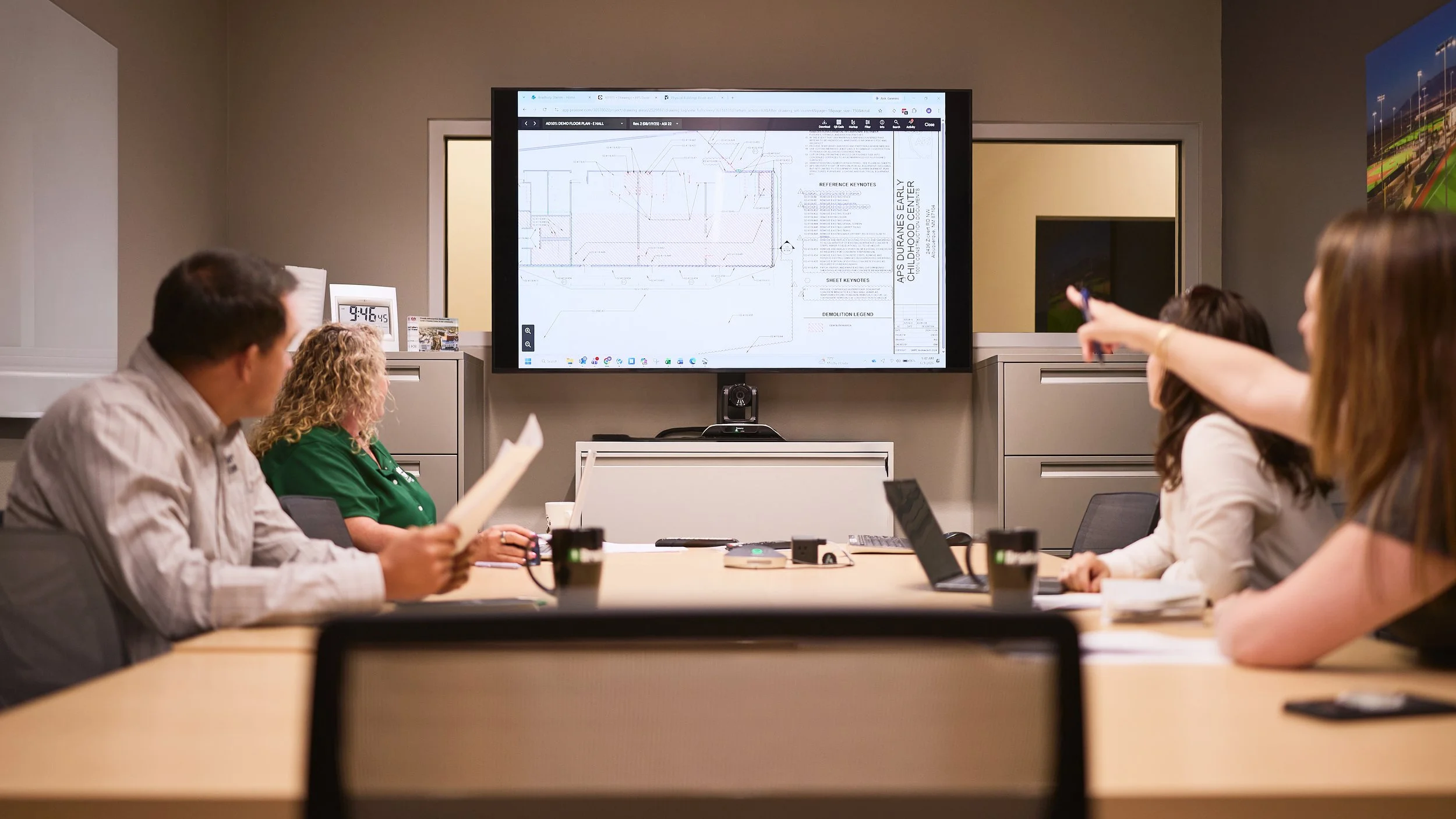 People seated at a conference table in a meeting room, looking at a large screen displaying technical plans or blueprints.