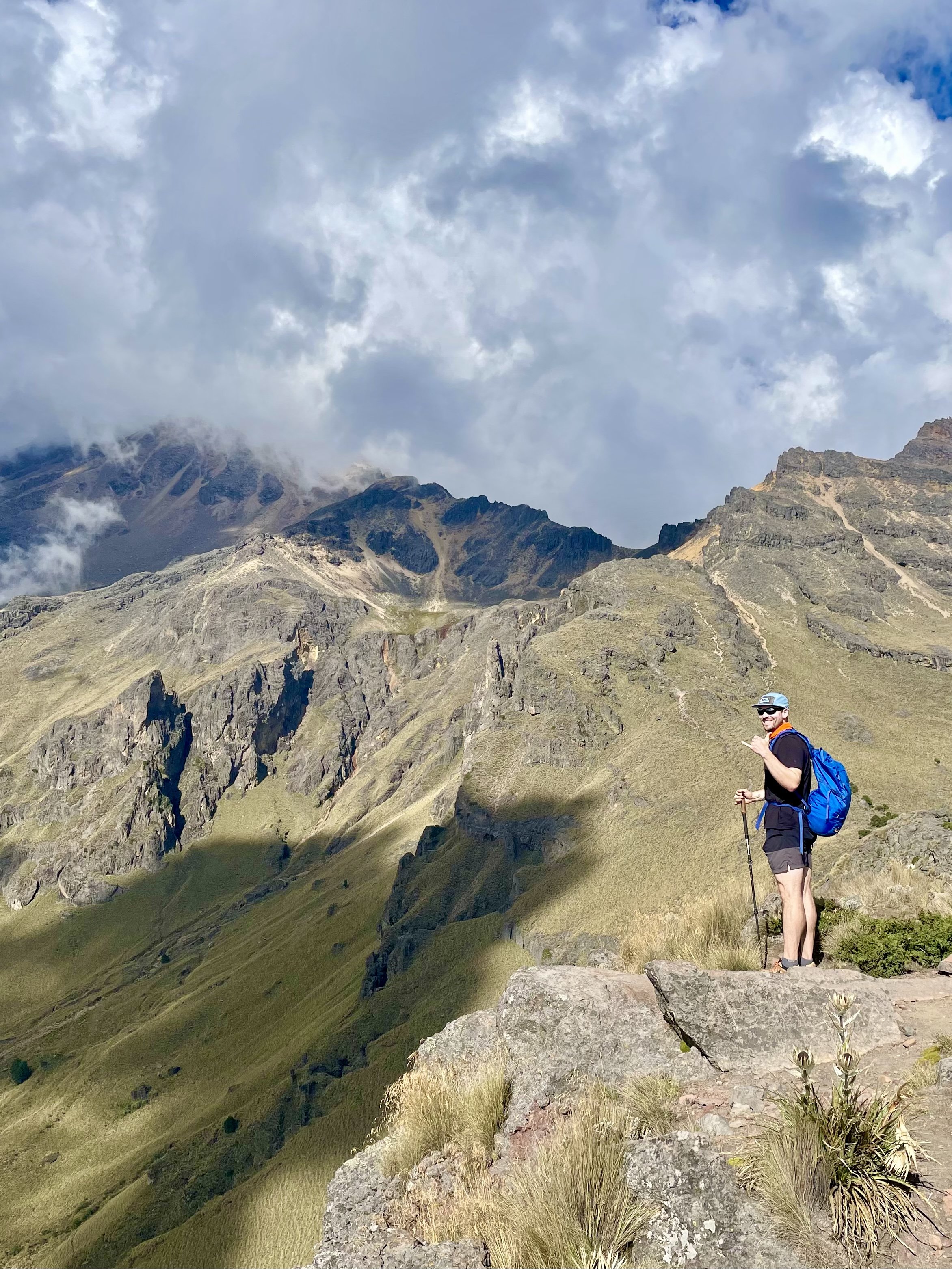 A man hiking on a mountain trail with a blue backpack, wearing a hat and sunglasses, holding a walking stick, surrounded by green grassy slopes and rocky outcroppings, with mountain peaks and cloudy sky in the background.
