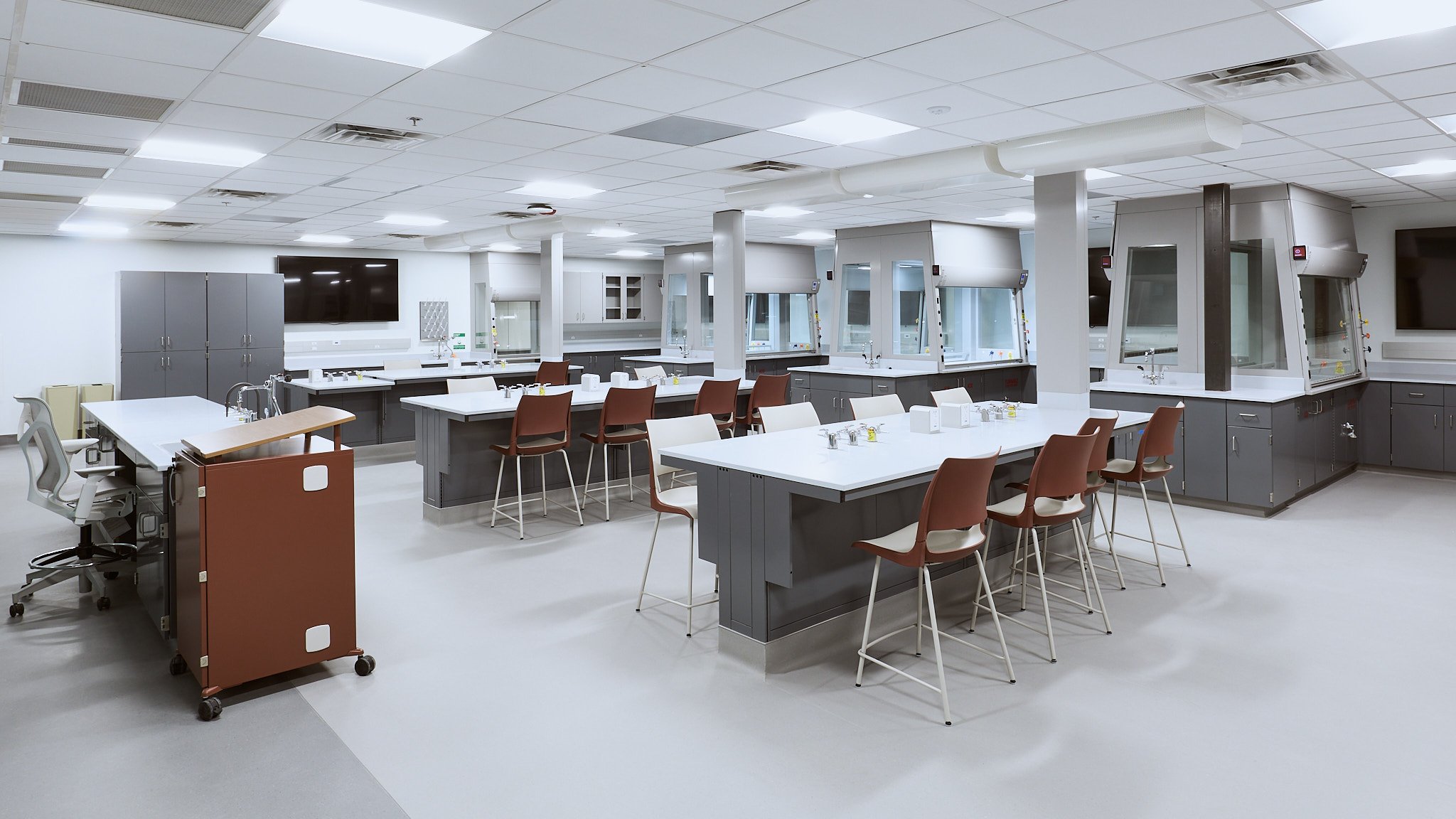 An empty laboratory with multiple workbenches, chairs, and scientific equipment, including fume hoods and sinks.