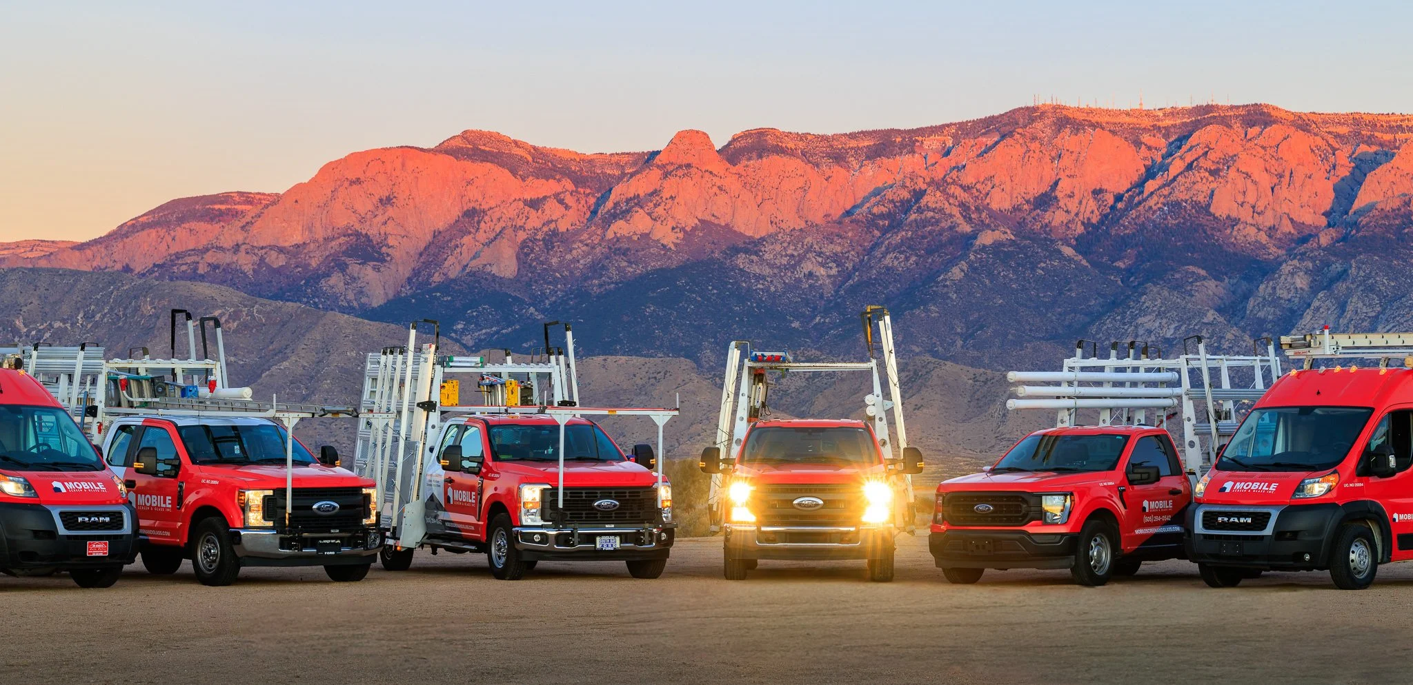A row of red service trucks with work equipment on the roofs, parked on a dirt surface with mountains in the background at sunset.