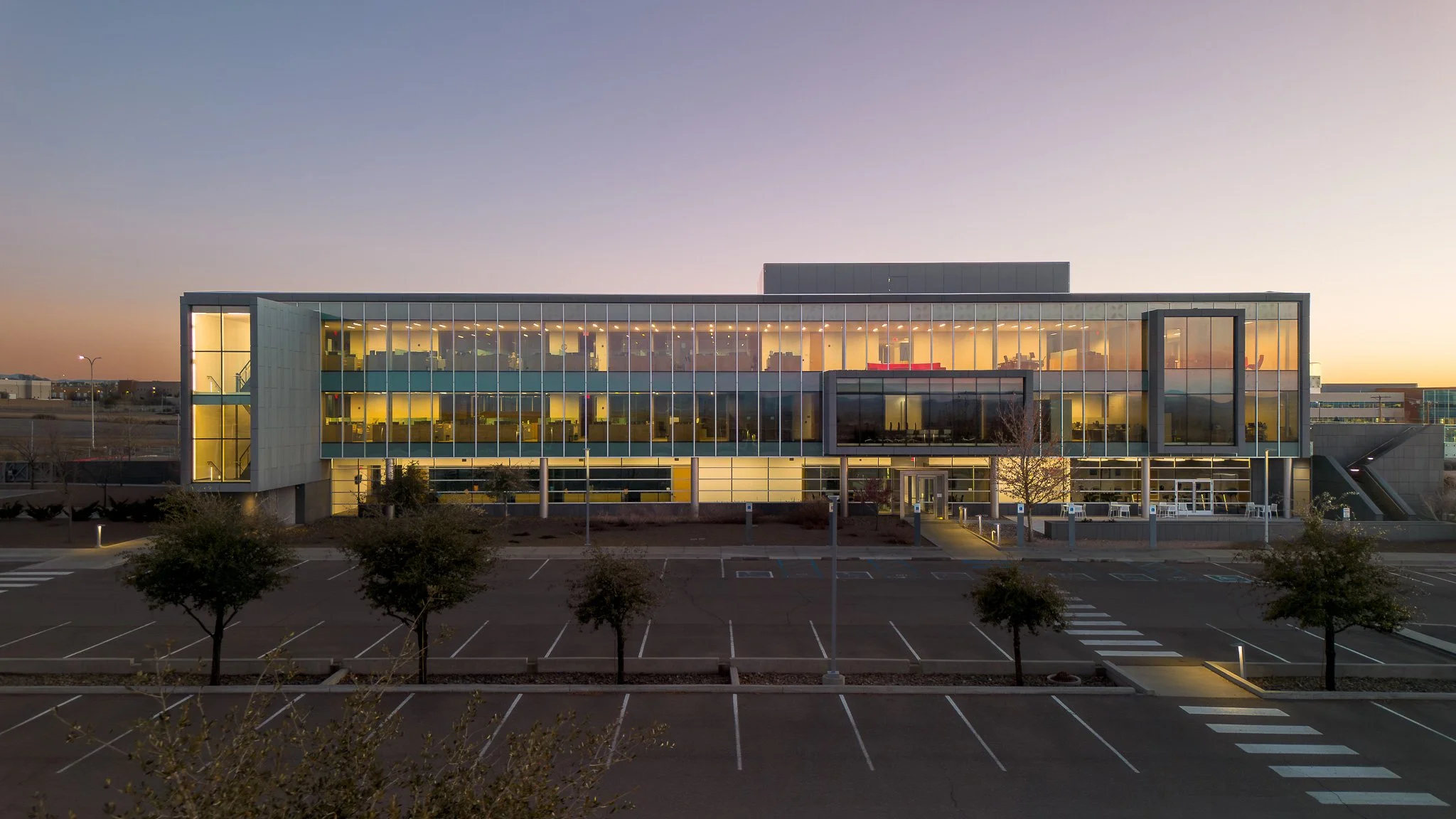 A modern multi-story office building with glass facade, lit from inside, at dusk with empty parking lot in foreground.