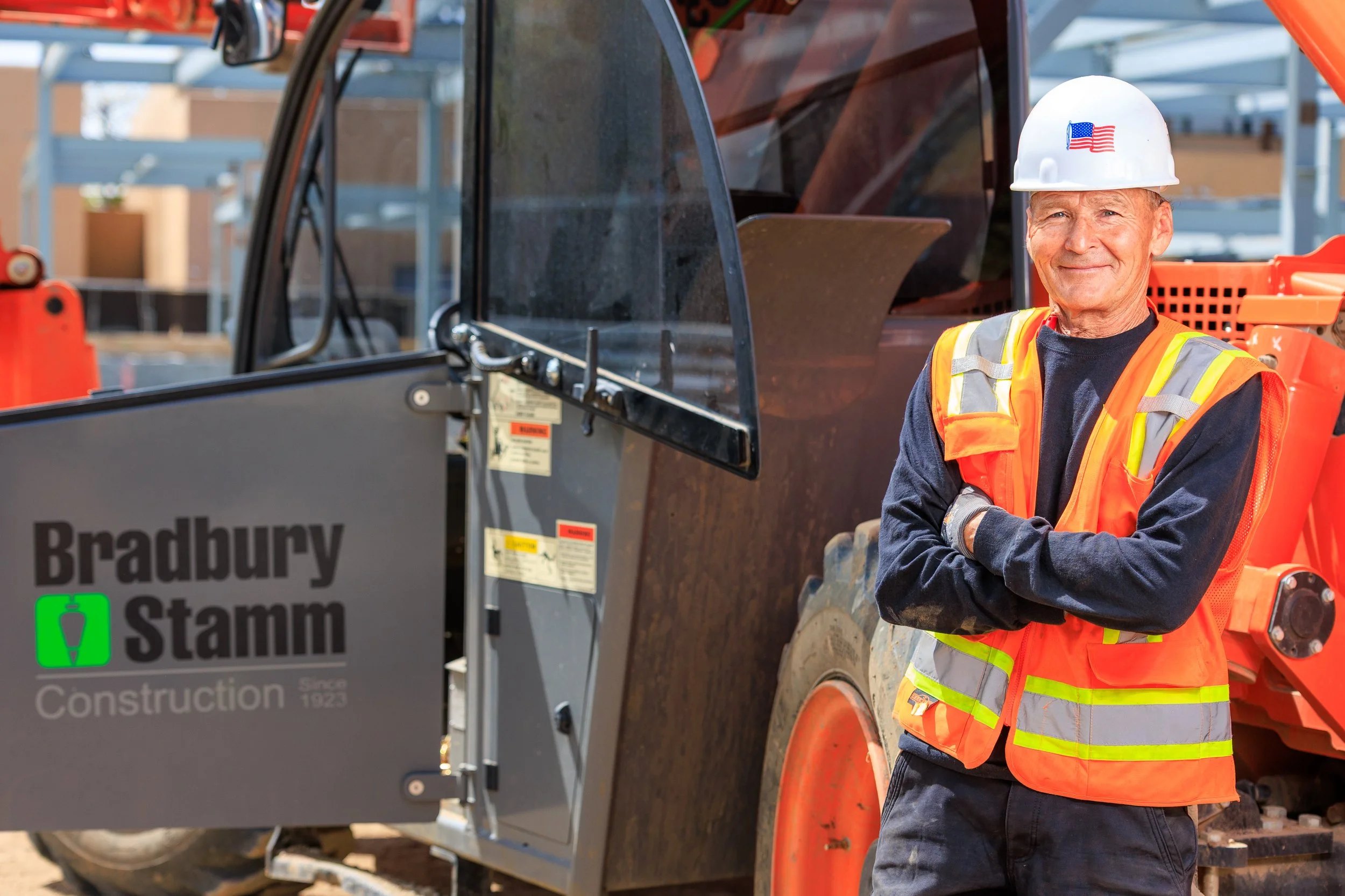 A construction worker with white hair smiling, wearing a white helmet with an American flag sticker, an orange safety vest with yellow reflective stripes, and standing with arms crossed in front of construction equipment at a building site.