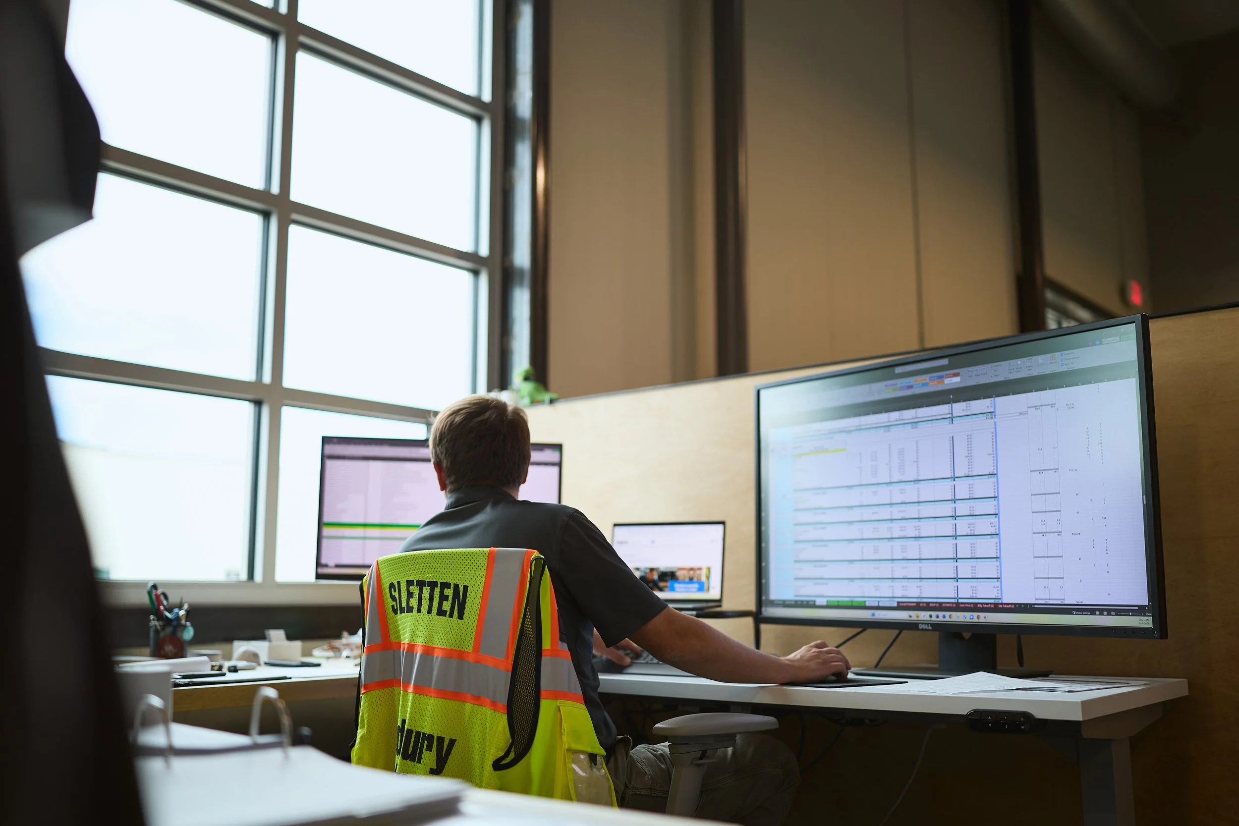An office worker sitting at a desk in front of a large computer monitor showing a detailed project schedule or timeline. The person is wearing a high-visibility safety vest with the label 'SLETTEN' on the back. The office has large windows letting in