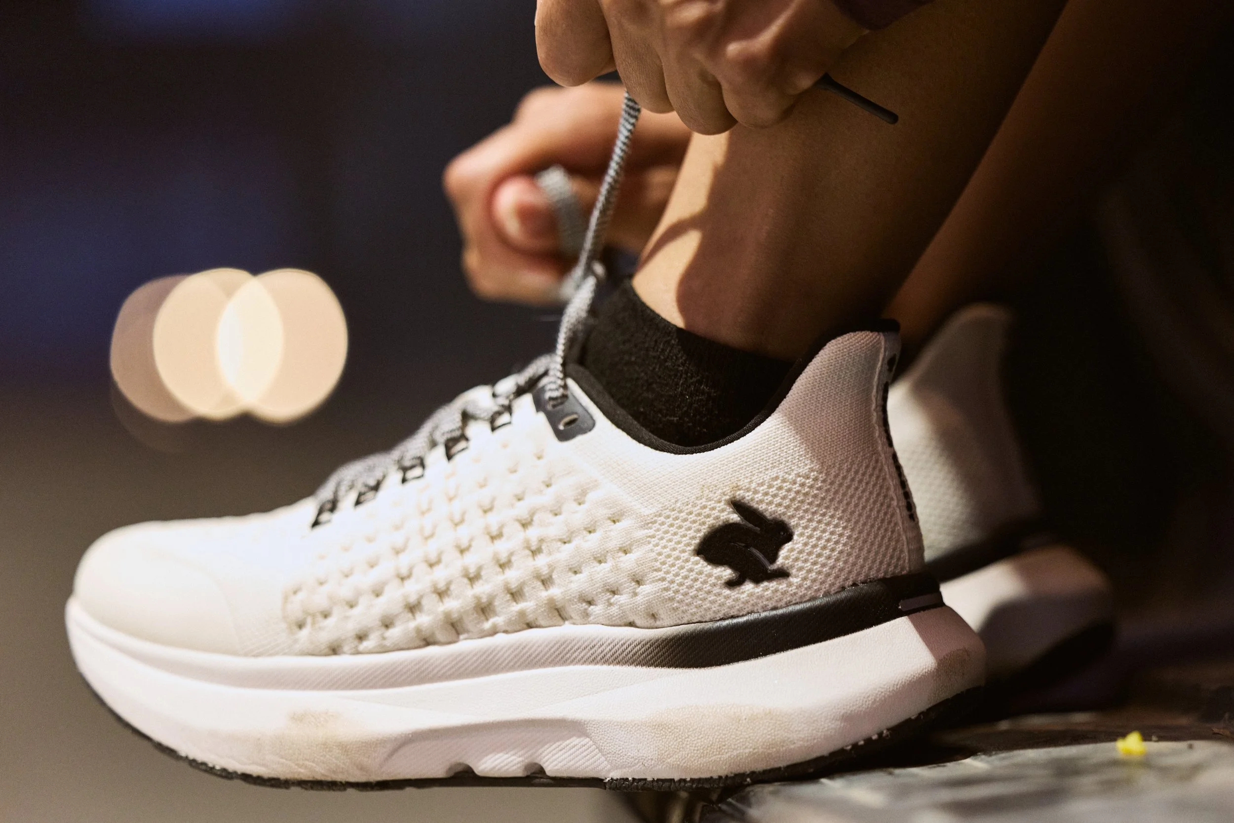 Close-up of a person tying shoelaces on a white athletic shoe with black accents, against a dark, blurred background.
