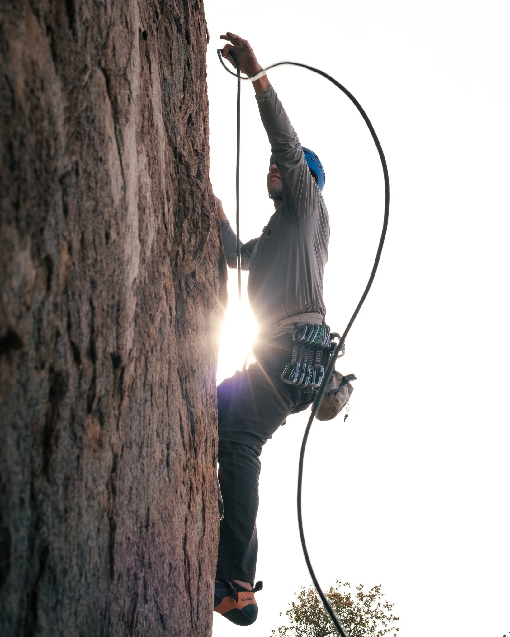 A person rock climbing on a steep wall with a harness and helmet, seen from below with the sun setting behind.
