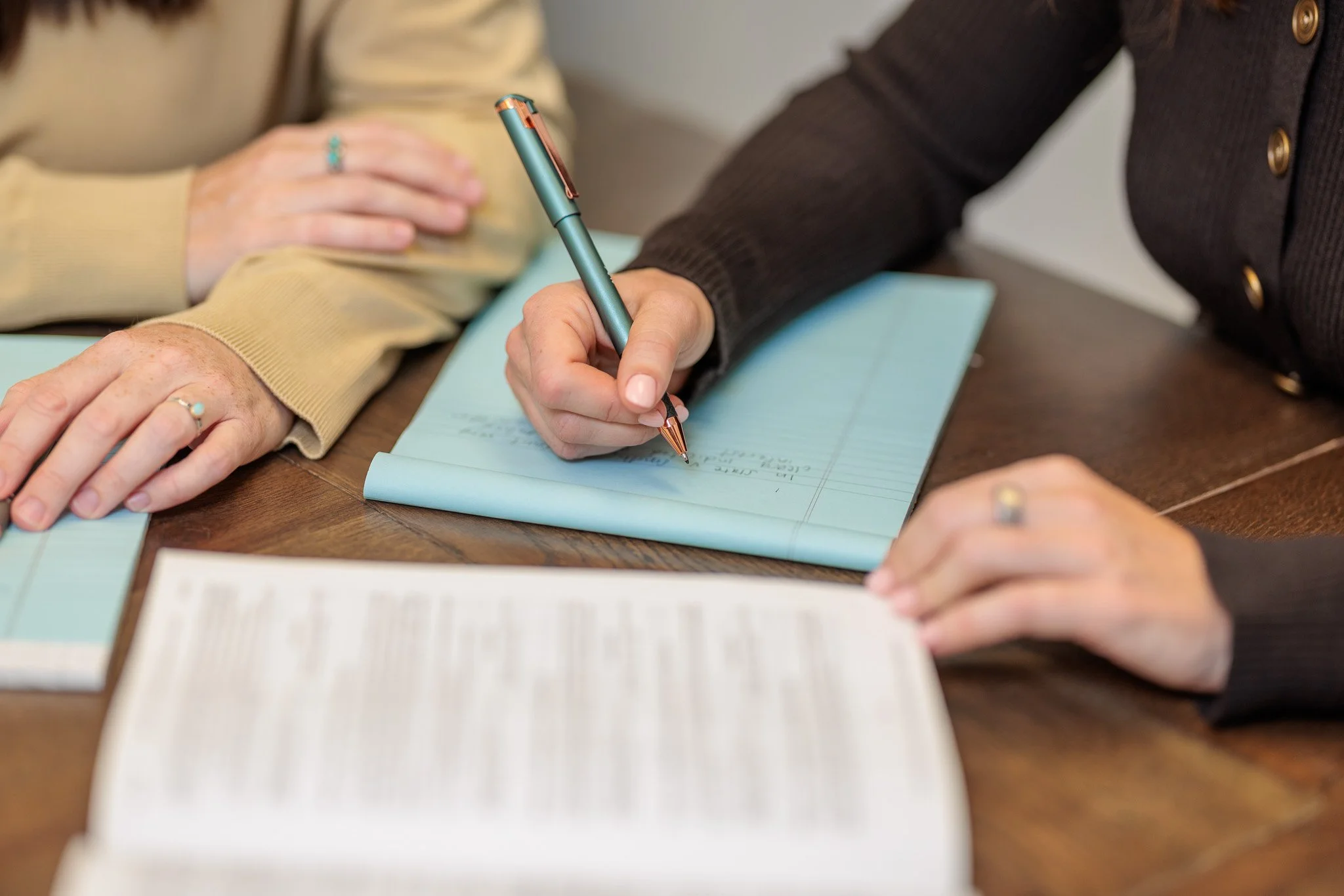 A person wearing a dark jacket is writing in a lined notebook with a teal and bronze fountain pen. Another person, wearing a cream-colored sweater, rests their hand on the table, which also has a book and papers.