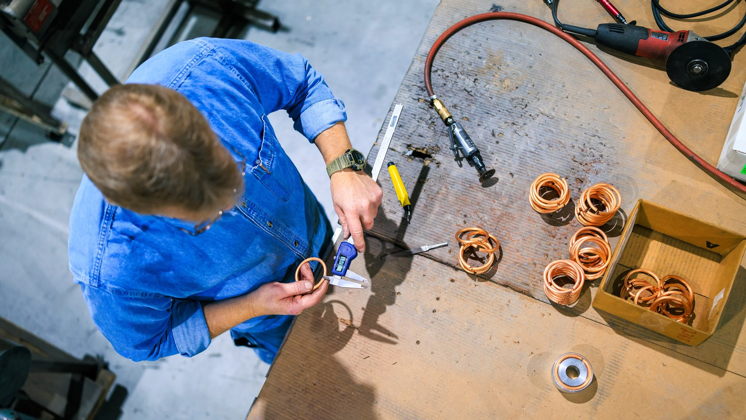 A person in a blue denim shirt working at a wooden table with copper coil components and tools related to electrical work.