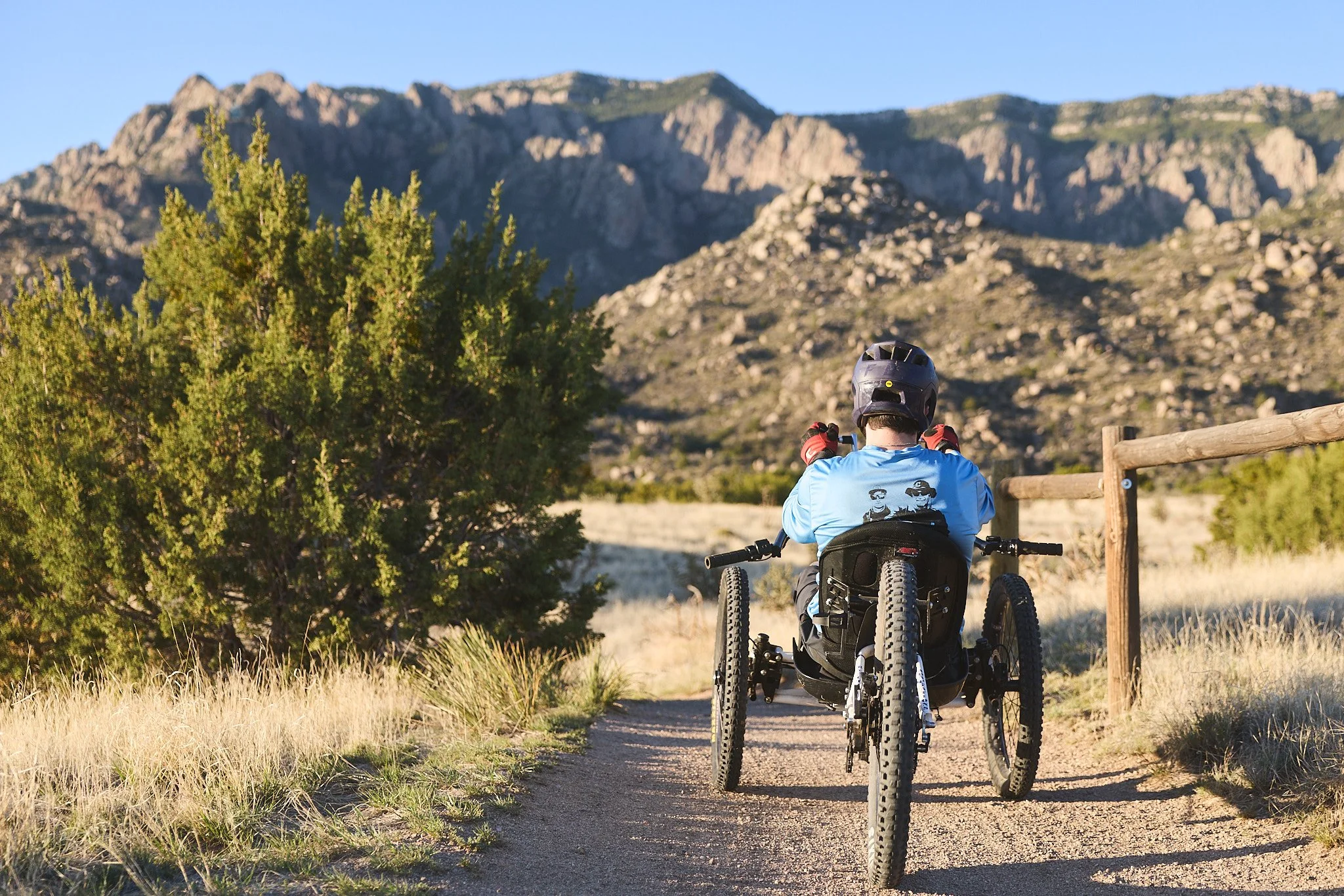 A person in a wheelchair riding an all-terrain bike along a dirt path in a mountainous area with green trees and rocky hills in the background.