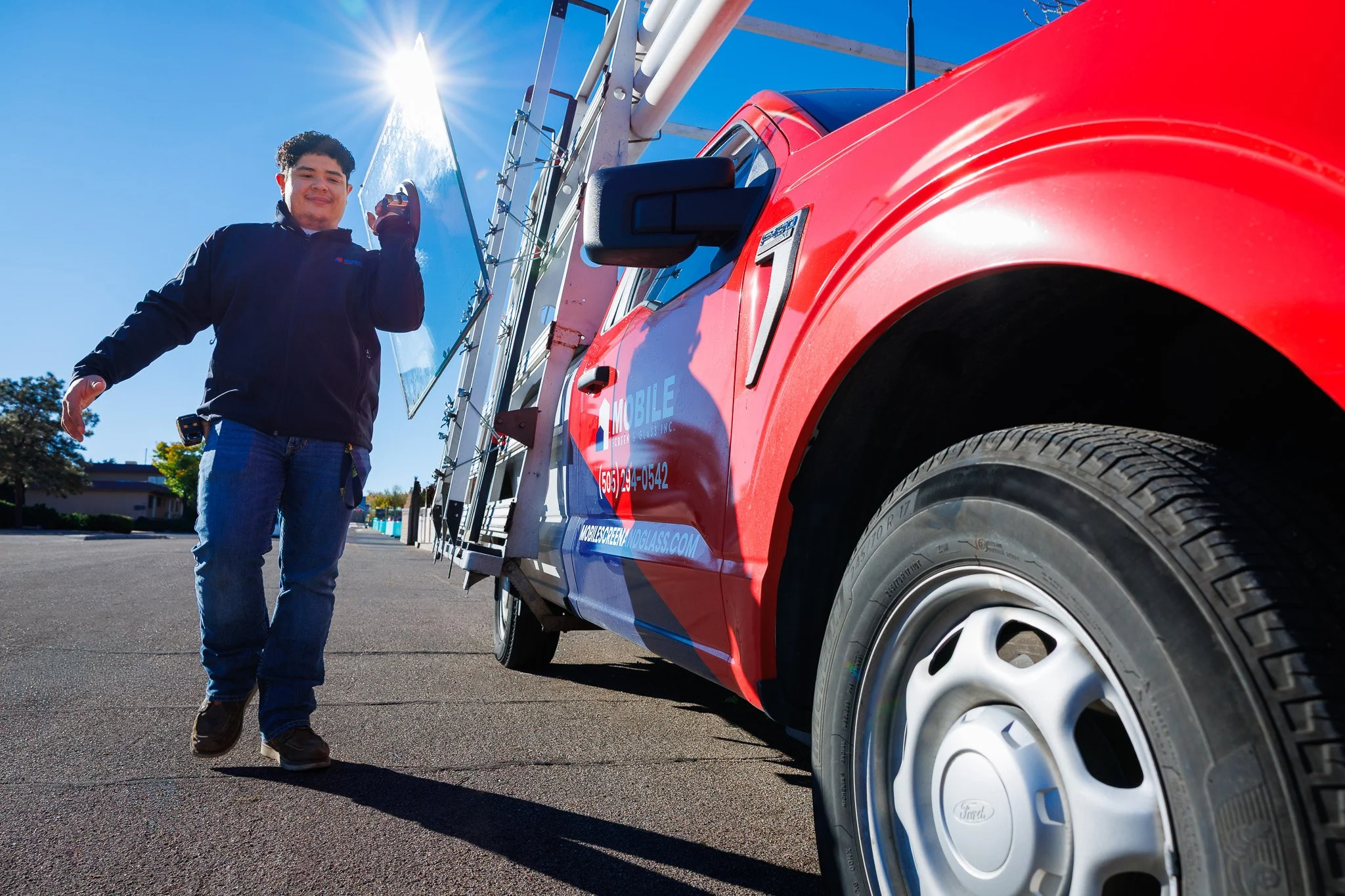 A young man with curly hair standing beside a red service truck on a sunny day, giving a thumbs-up gesture, with a ladder and a solar panel on the truck.