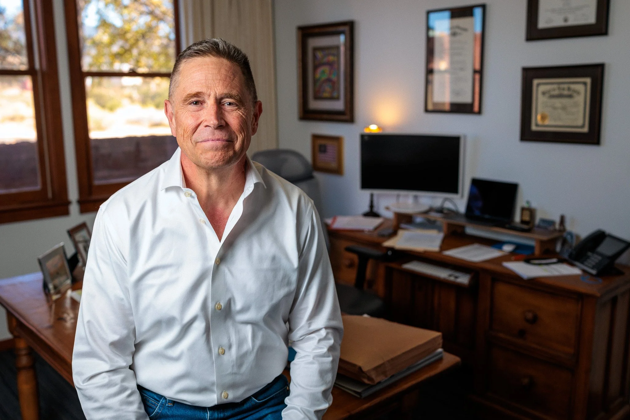 A middle-aged man with short hair wearing a white collared shirt, standing in an office with framed artwork and certificates on the wall behind him. There is a large window to his left bringing in natural light.