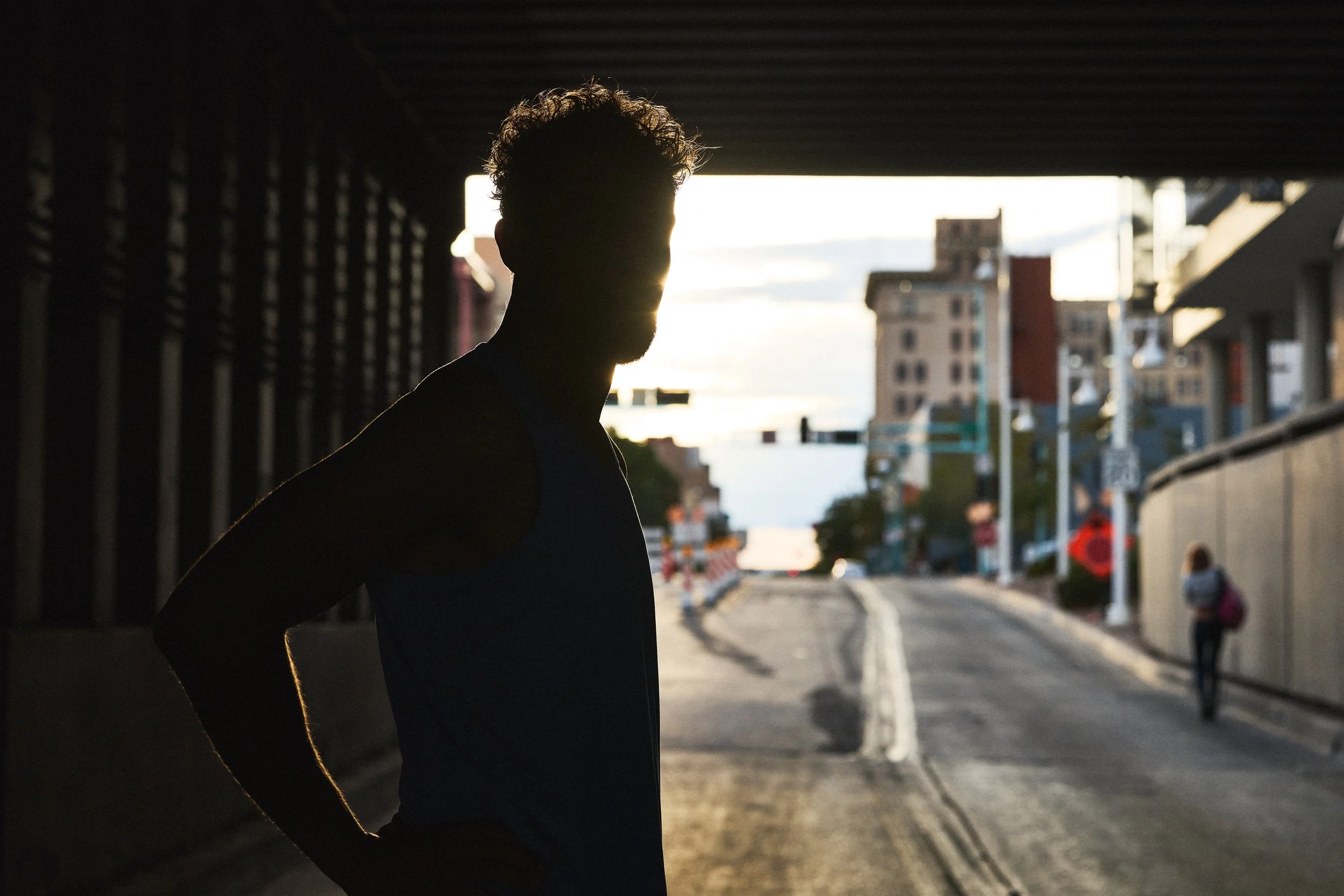 Silhouette of a person with curly hair under an urban overpass at sunset, with city buildings and a street in the background.