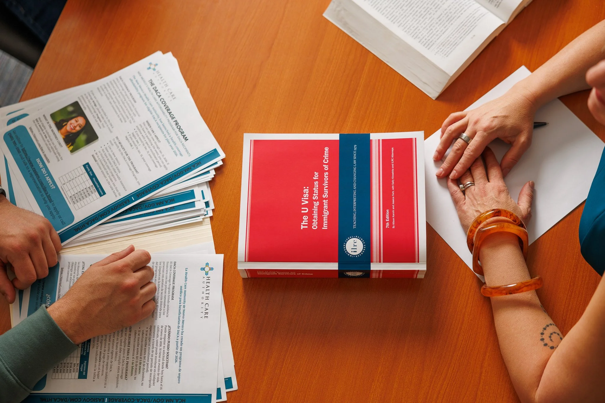 Hands on a wooden table with brochures, a book, and a pen. One person has a silver ring and another has a black tattoo on their wrist.