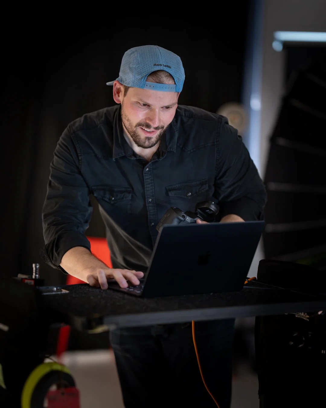 Man in a black shirt and blue baseball cap working on a laptop in a dimly lit room.