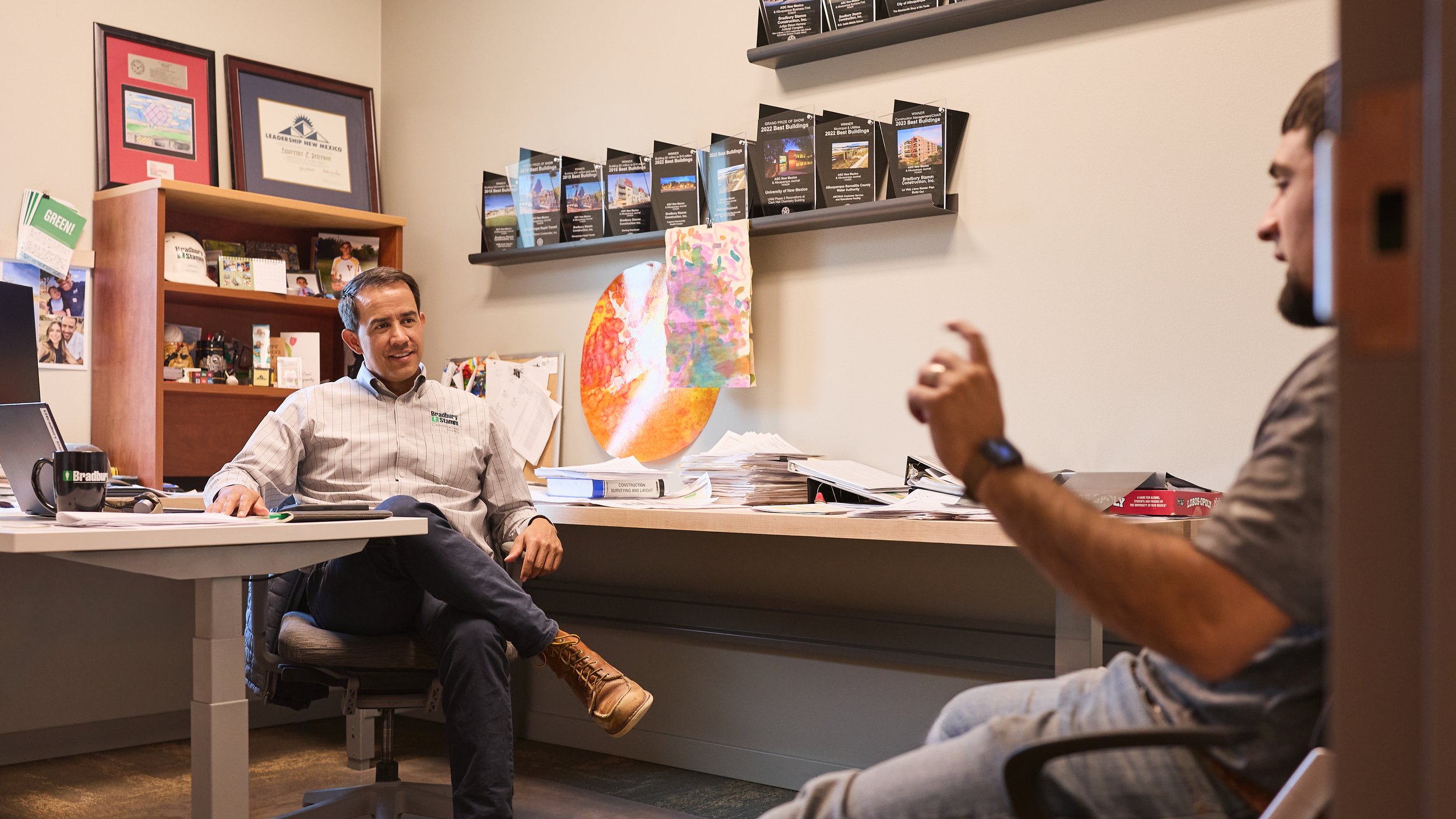Two men having a conversation in an office. One man is sitting behind a cluttered desk and smiling, while the other is seated across from him, gesturing with his hand.