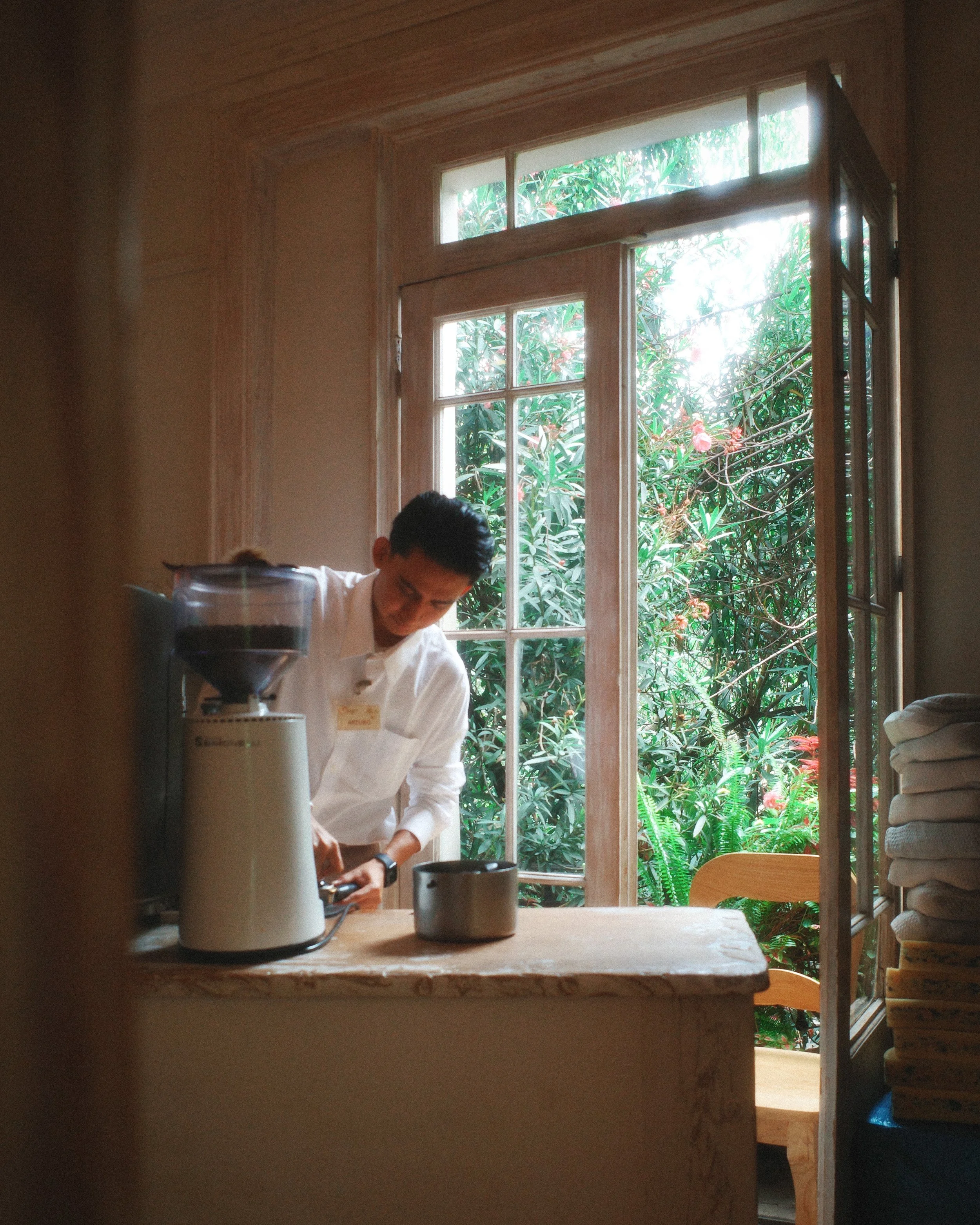 Person in white apron working in a kitchen with a large window showing green foliage outside.
