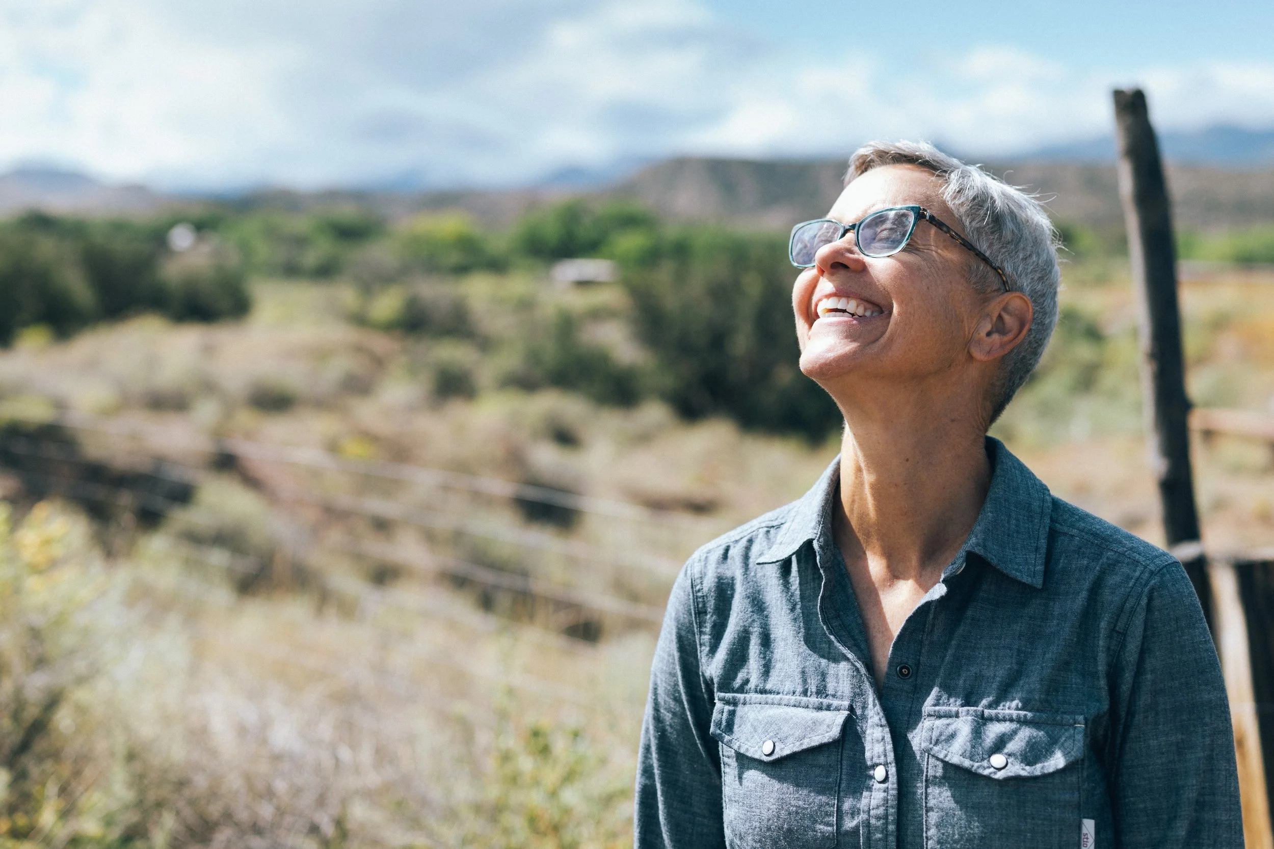 Smiling elderly man with gray hair and glasses looking up outdoors in a rural setting.