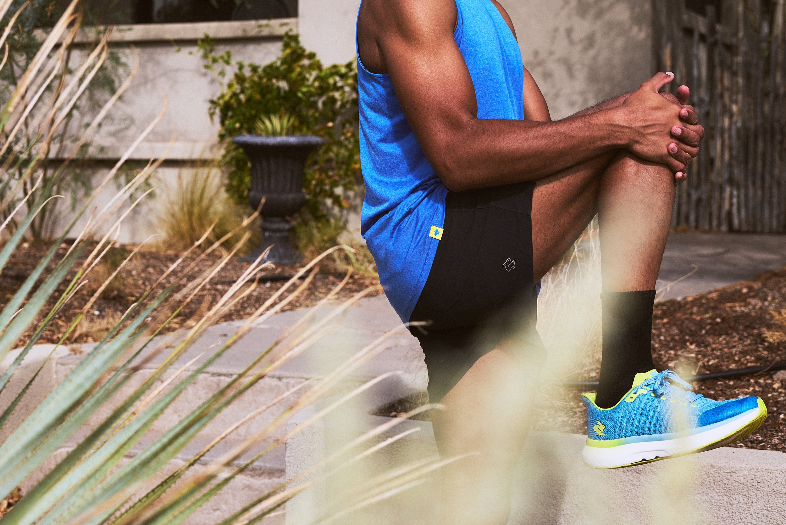 A man stretching outdoors wearing a blue tank top, black shorts, and blue running shoes with yellow accents on a concrete sidewalk, with plants and a wooden fence in the background.