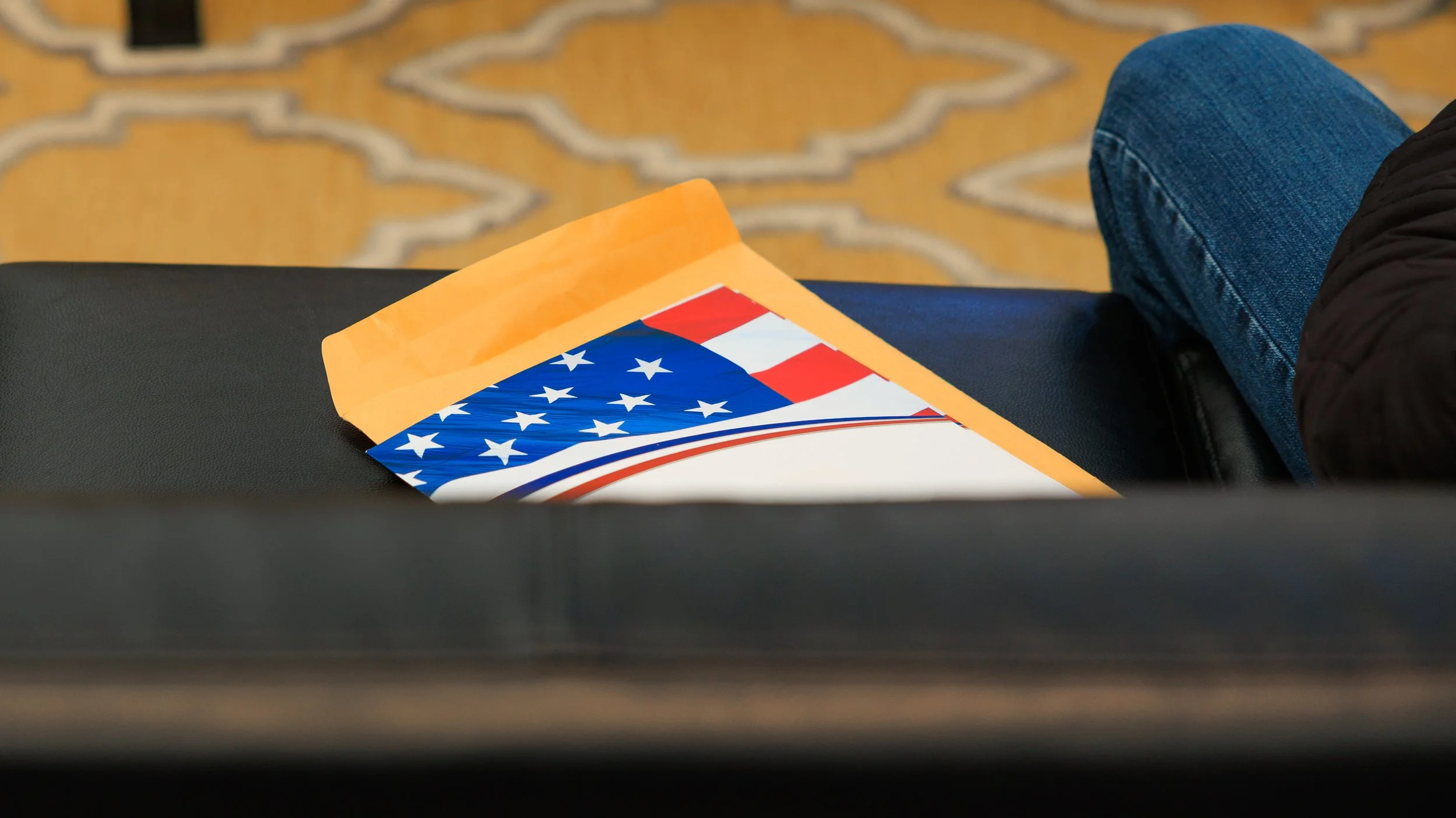 Part of an American flag inside a yellow folder on a black surface with a person wearing blue jeans and a black jacket sitting nearby, and a patterned carpet in the background.