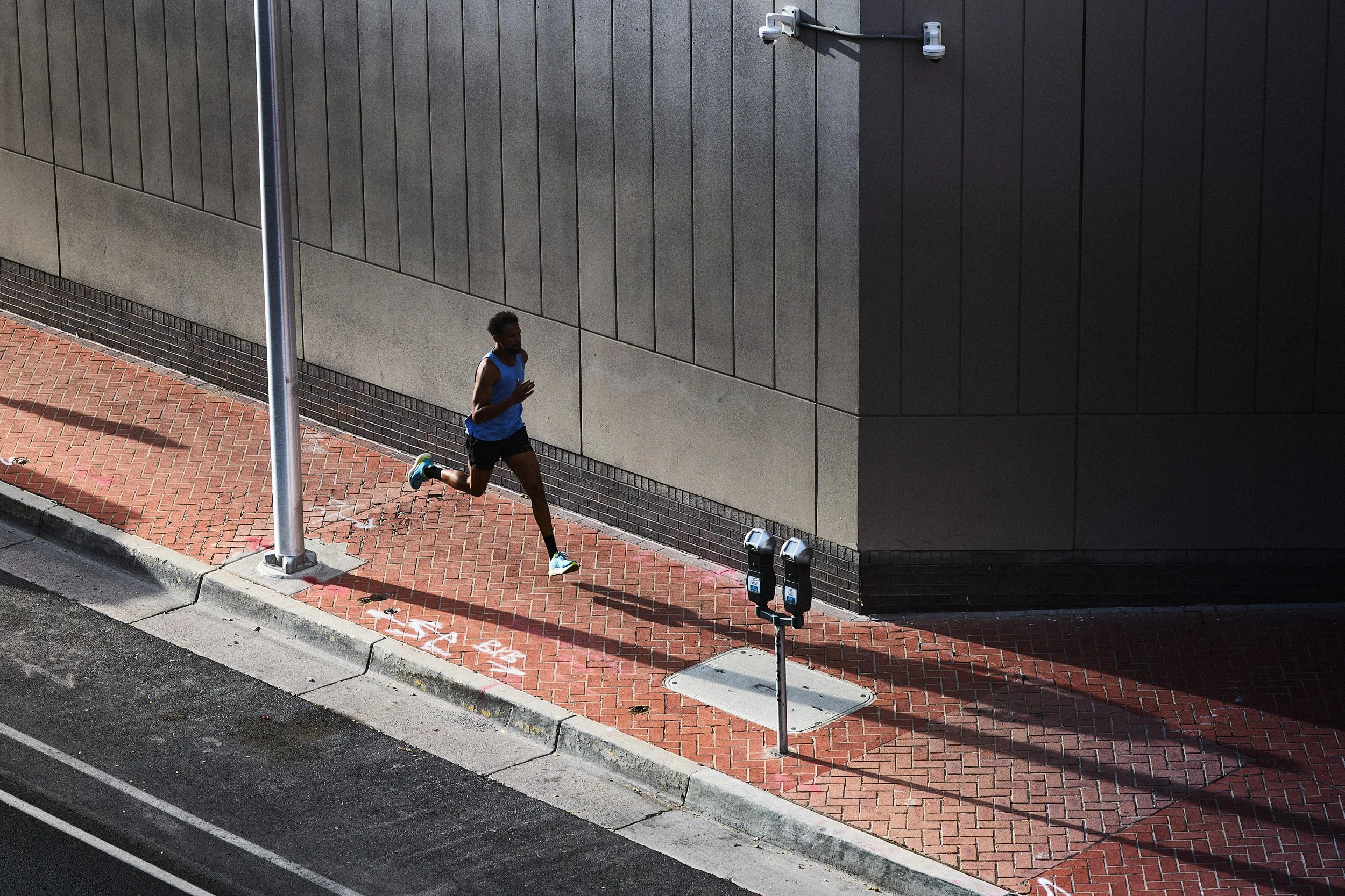 A person jogging on a red brick sidewalk next to a large gray building with a black wall. There is a lamppost and parking meters nearby.