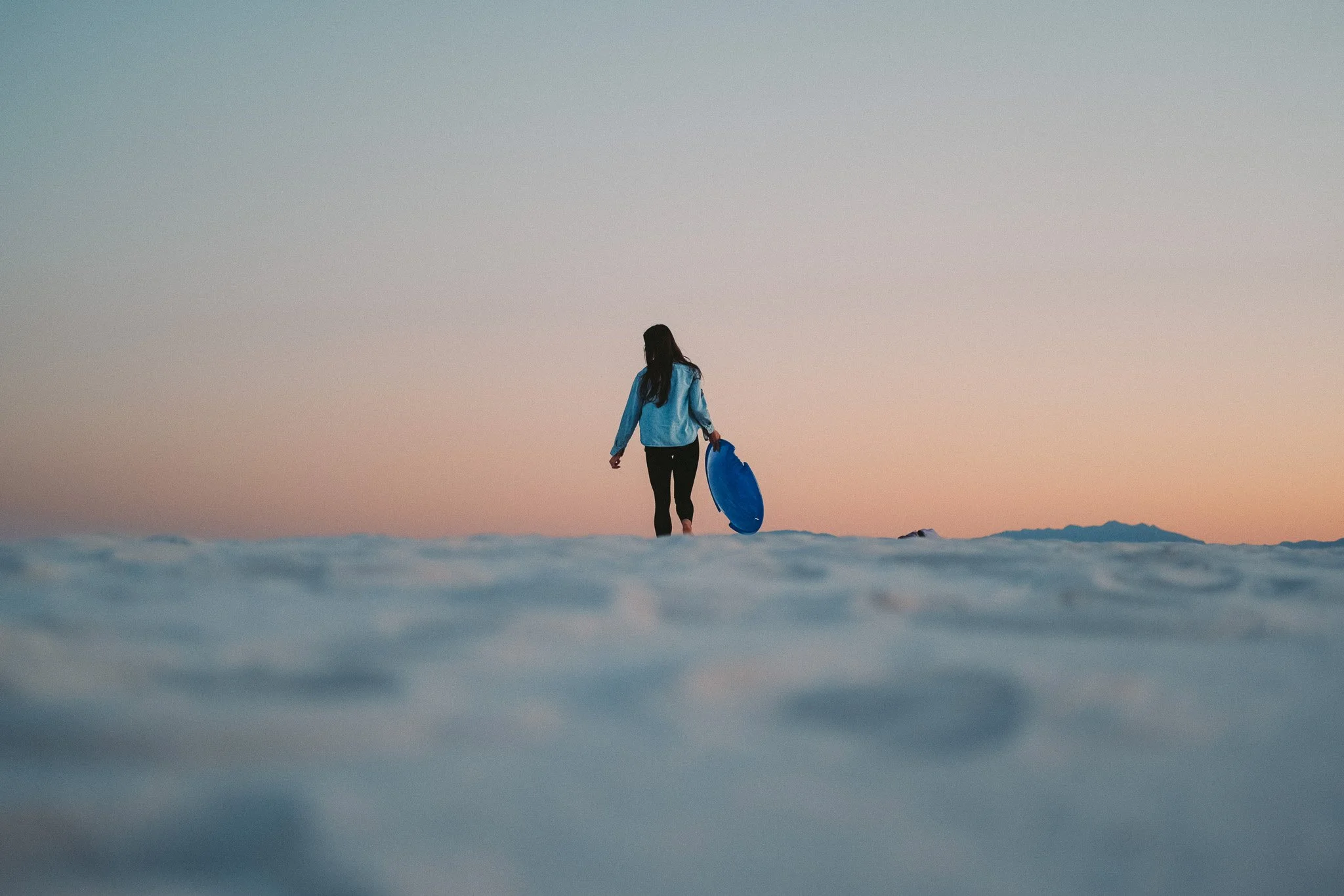 A woman in a blue shirt holding a blue boogie board walking on the beach during sunset.