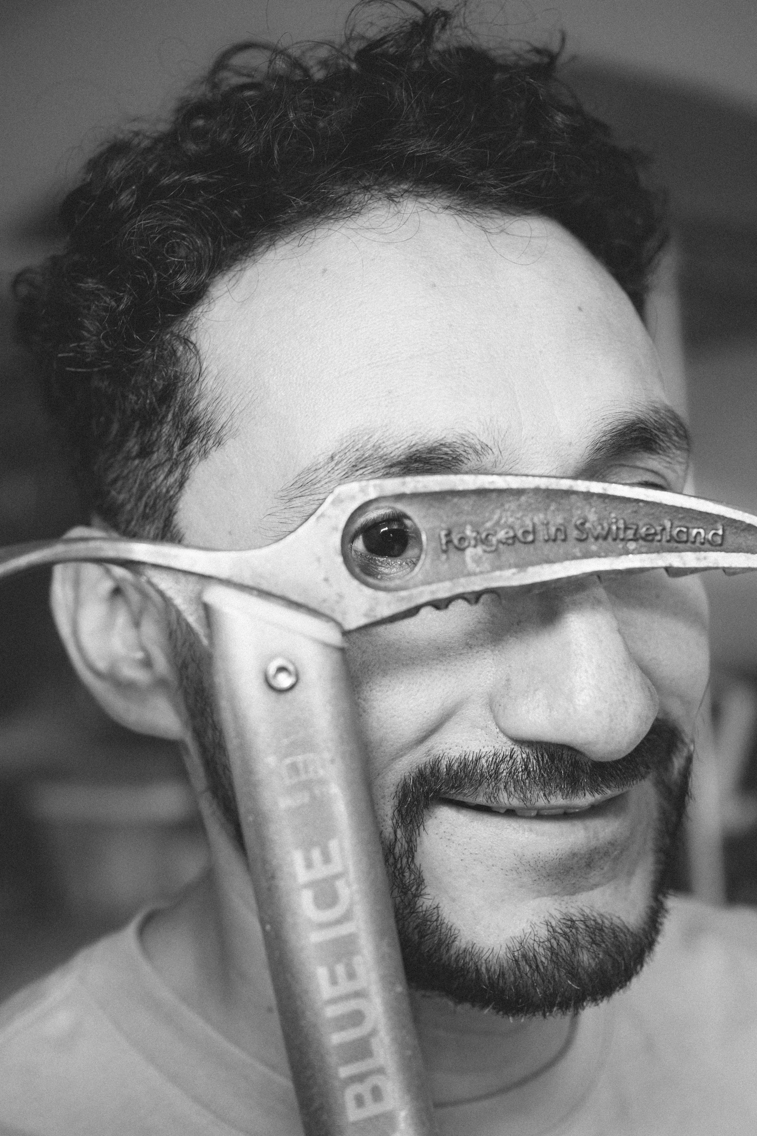 Black and white close-up photo of a man with dark curly hair and a beard, smiling, wearing protective goggles with the wording 'Made in Switzerland.' The goggles are positioned over his eyes, and he is looking slightly down.
