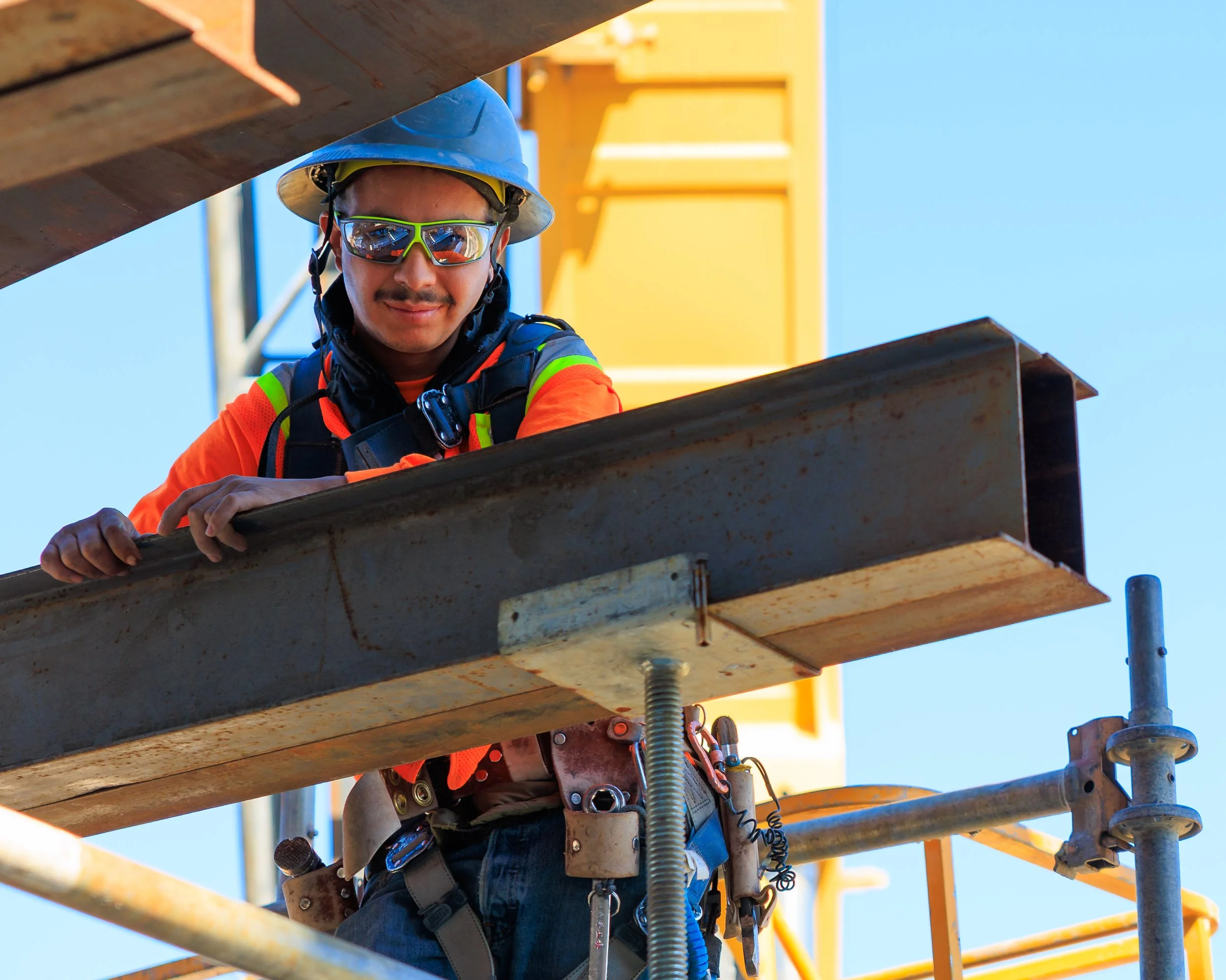 A construction worker wearing a blue safety helmet, reflective sunglasses, and an orange safety vest, is working on a metal structure at a construction site. The worker is focused and smiling, with a blue sky in the background and yellow construction