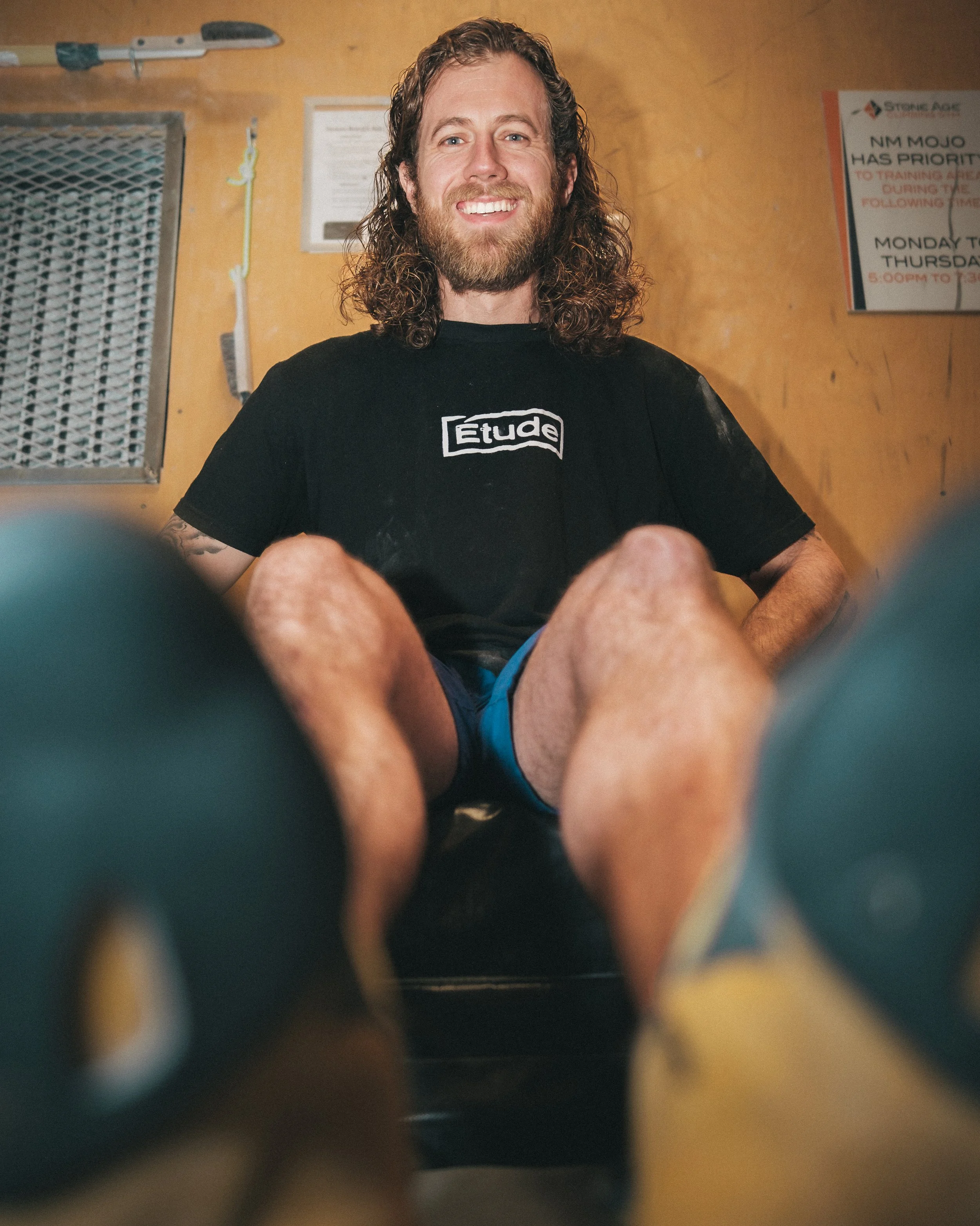 A smiling man with long curly hair and a beard, wearing a black T-shirt with 'Étude' written on it, is sitting in a room with yellowish walls. Behind him, there are some signs and a toolbox hanging on the wall.
