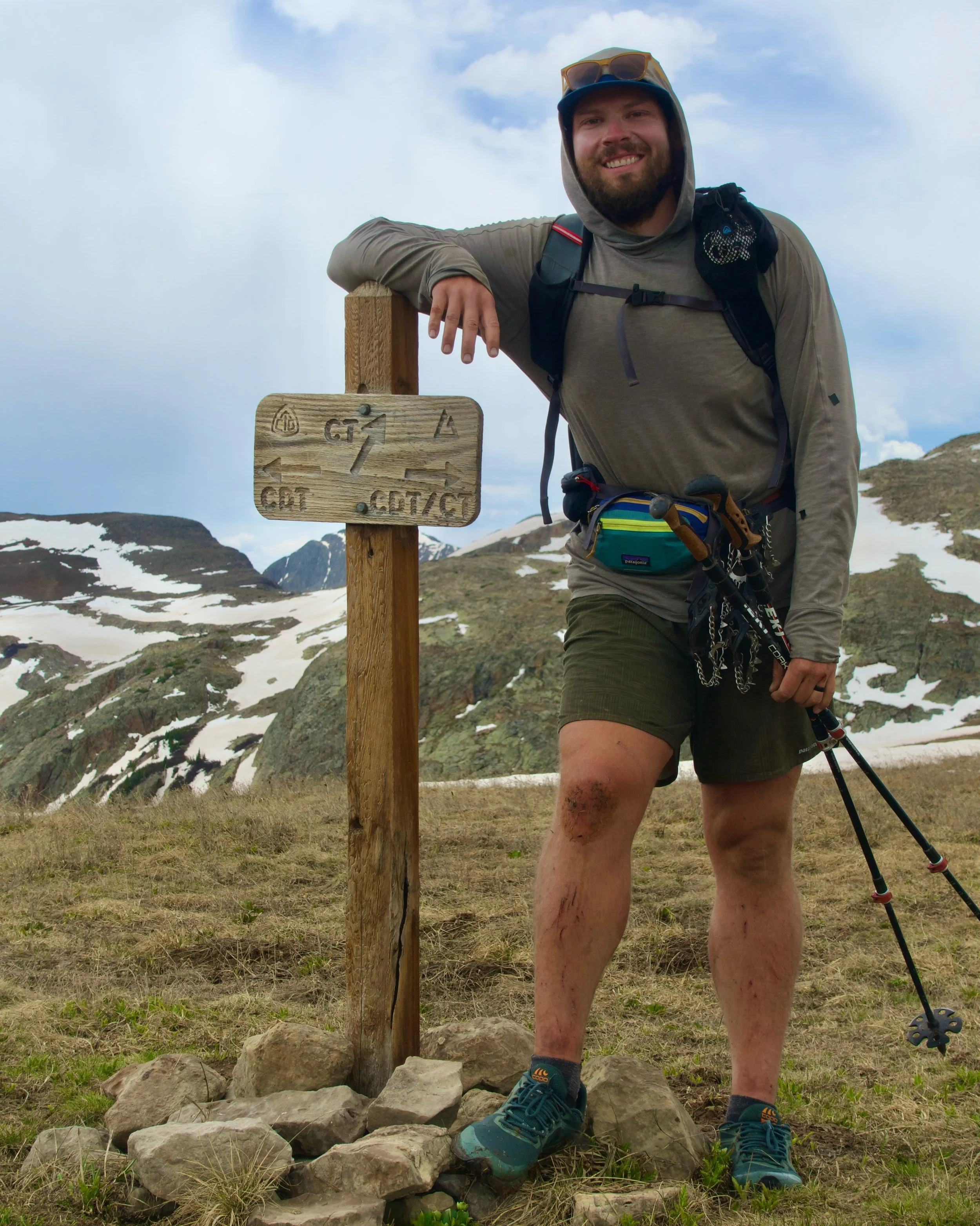 A smiling man with a beard wearing a hoodie, sunglasses on his head, and hiking gear, resting on a wooden signpost on a mountain trail with snow-capped mountains in the background.