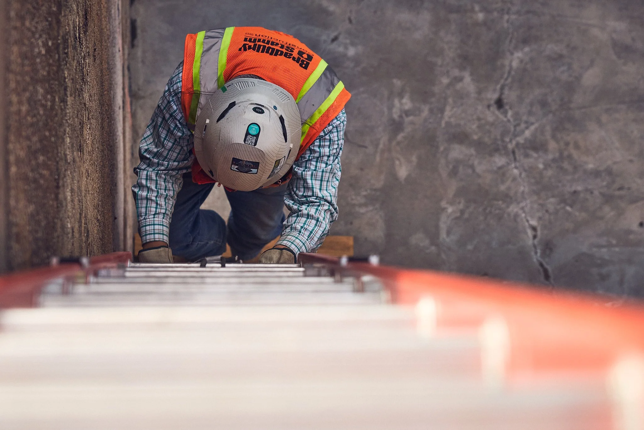 A construction worker wearing a hard hat and a reflective safety vest is climbing a ladder on a construction site. The worker is on their hands and knees, looking down, with a concrete wall to the left and a textured gray wall behind.