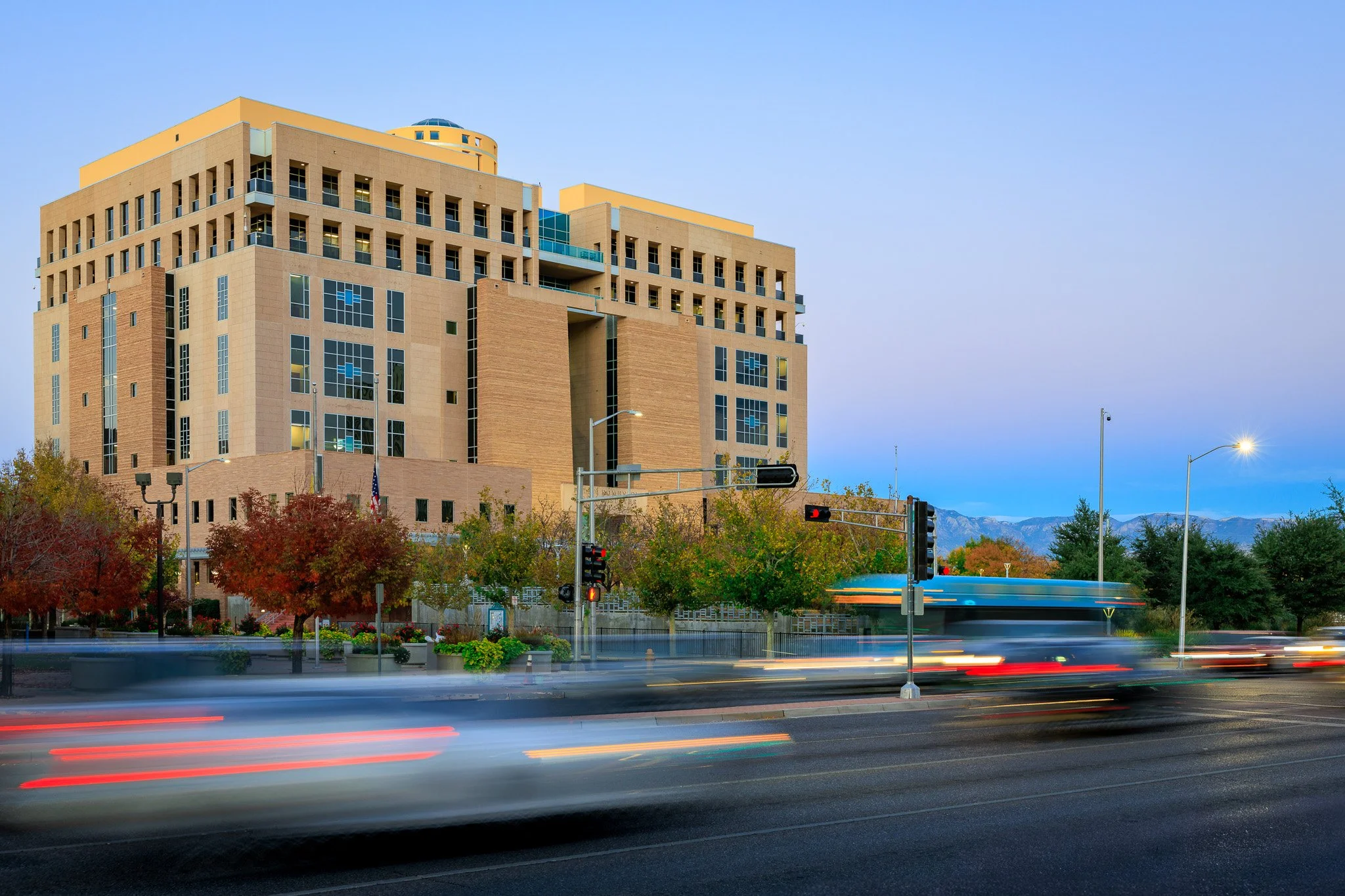 A large modern multi-story building with beige and brown exterior, multiple windows, and a rounded top section. In the foreground, a busy street with moving vehicles creates light trails. There are trees with fall foliage and a distant mountain range