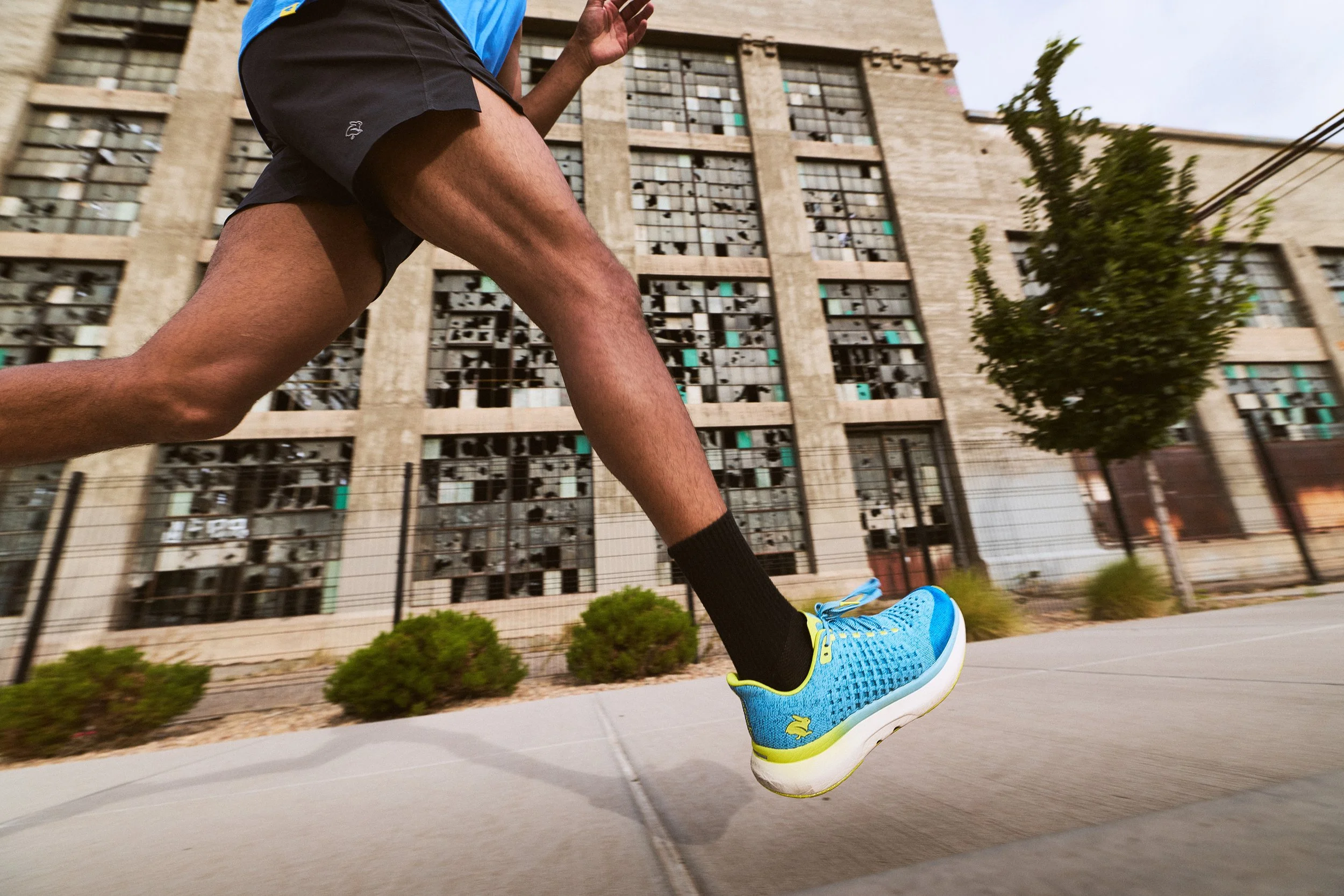Close-up of a person running outdoors on a sidewalk, wearing blue running shoes, black socks, shorts, and a blue shirt, with a building and a small tree in the background.