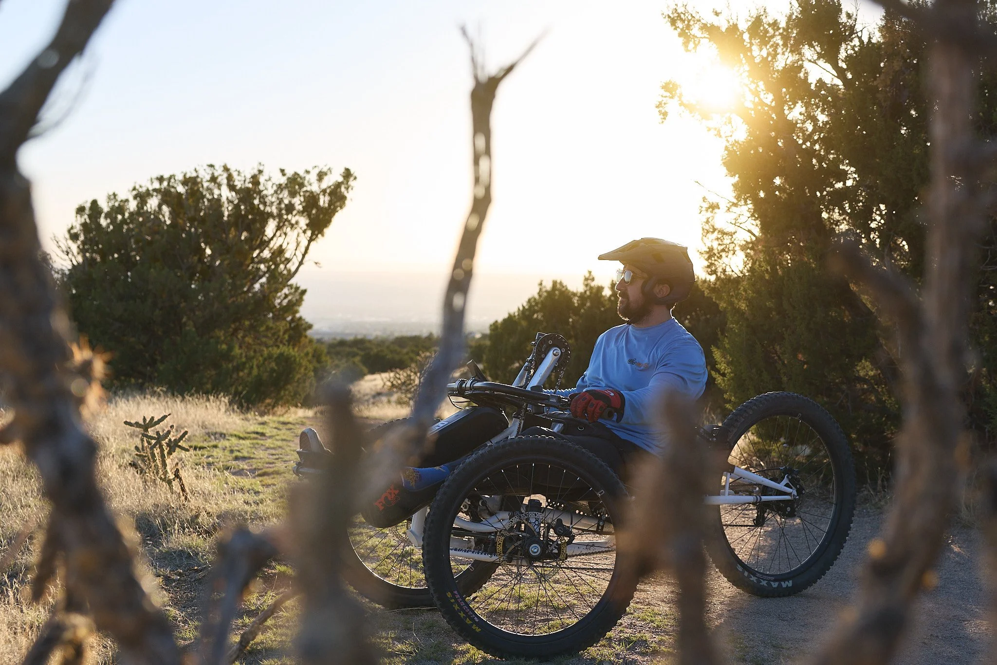 A man with a beard and glasses, wearing a helmet, sitting on a mountain bike in a natural outdoor setting during sunset or sunrise, framed by tree branches and foliage.