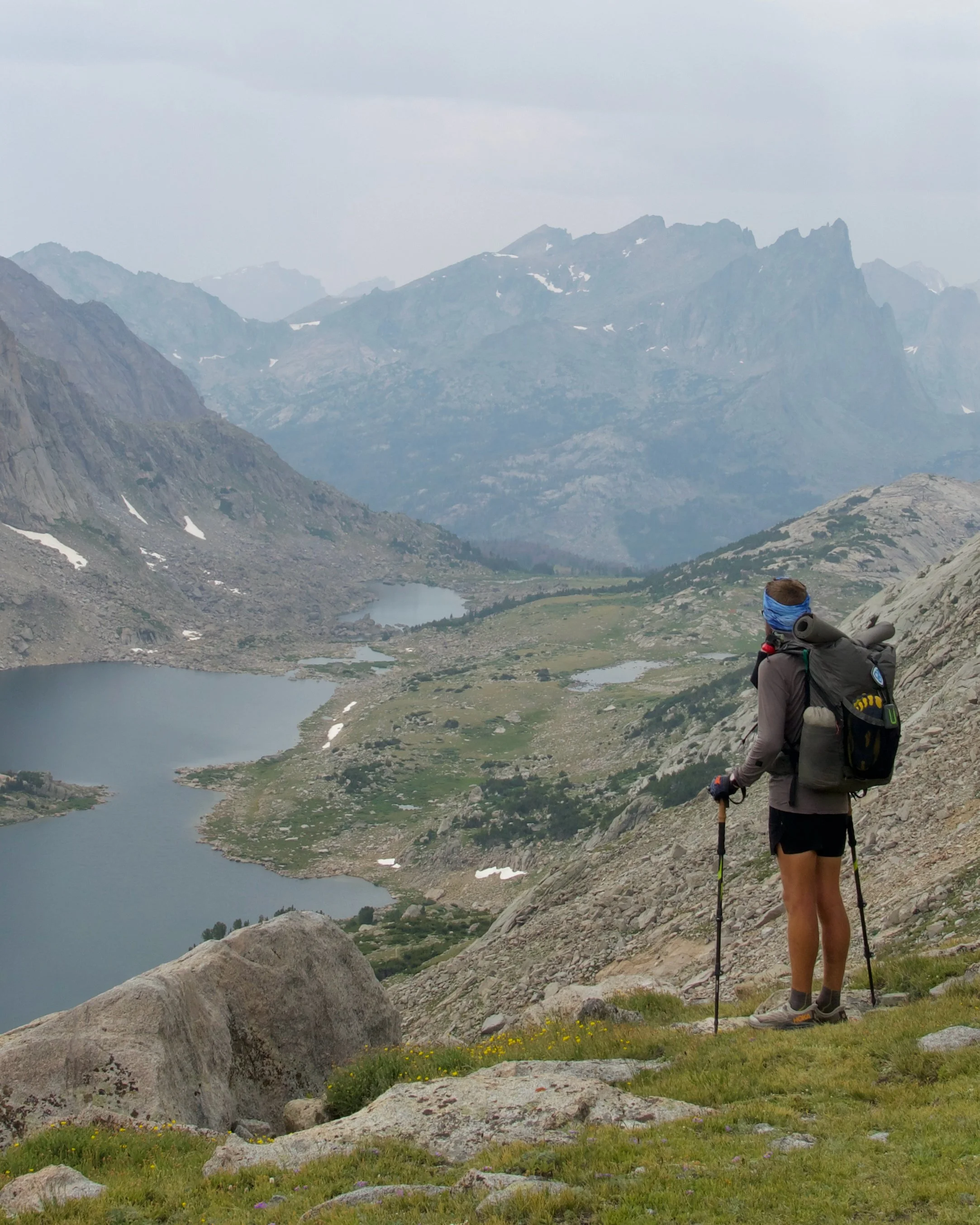 Hiker in outdoor mountain landscape with lakes, rocky terrain, and snow-capped peaks in the distance.