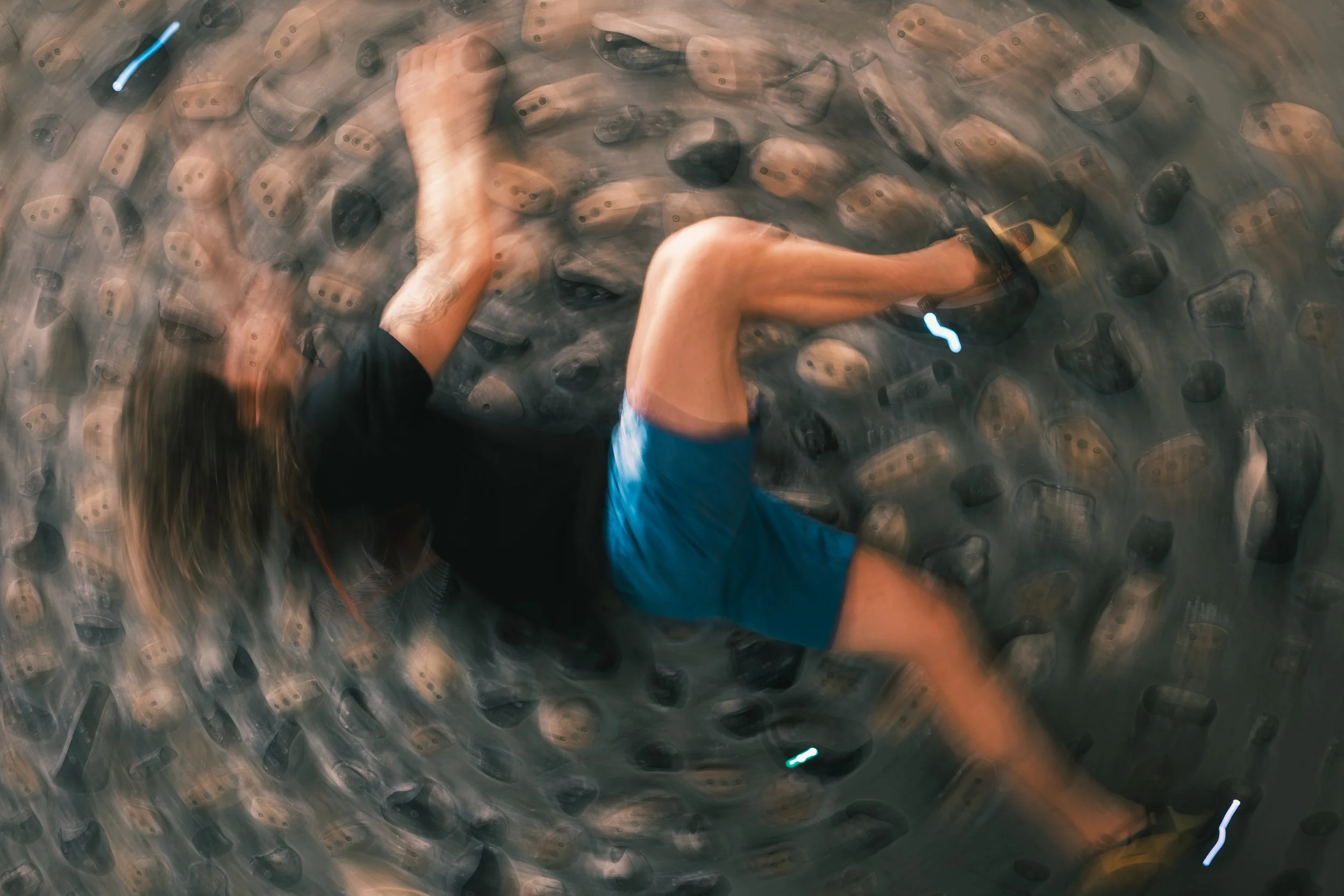 Person climbing an indoor rock wall, wearing a blue shirt and harness, moving upward.