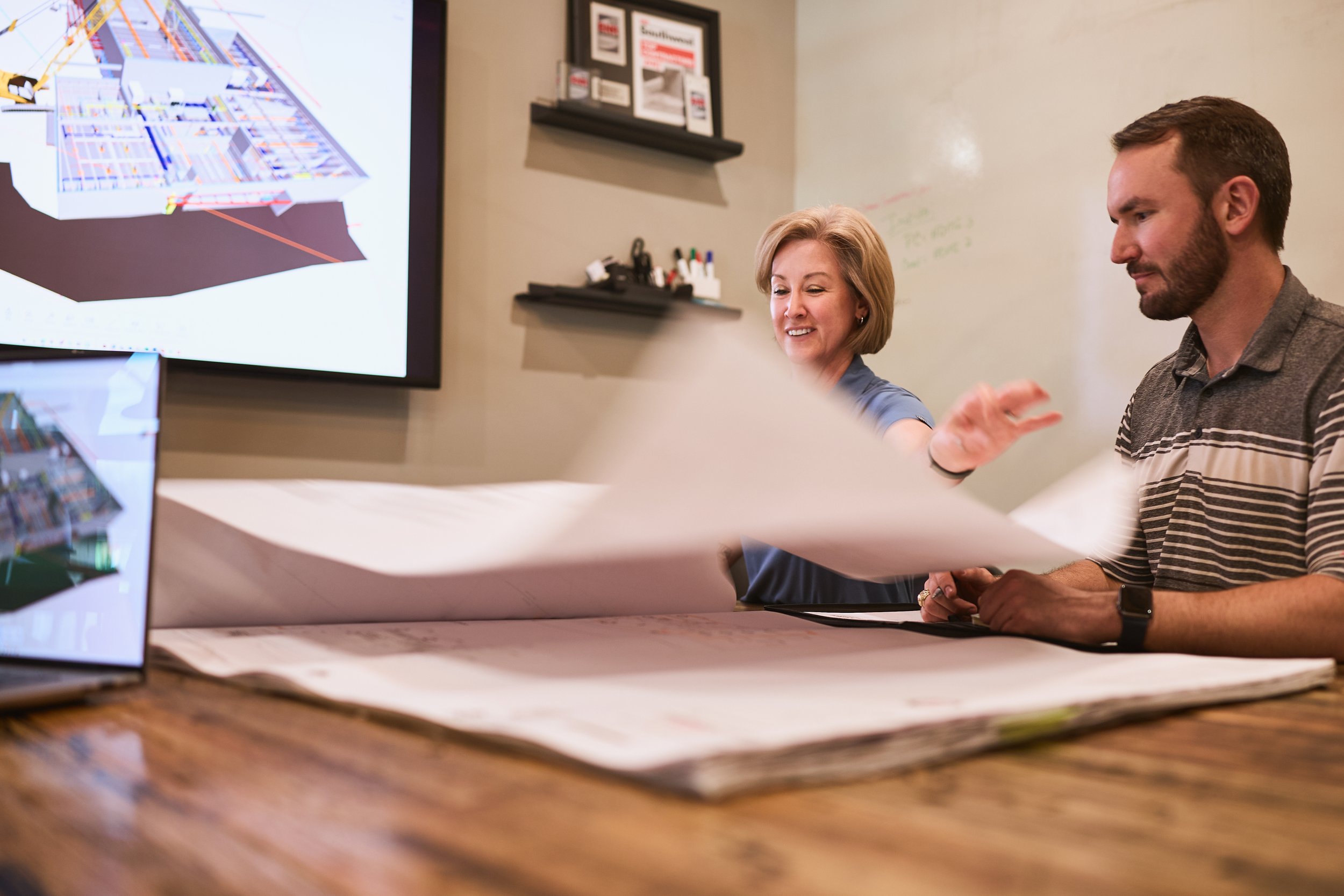 Two people, a woman with blonde hair and a man with short brown hair and a beard, are sitting at a table in front of a large screen, discussing a blue and white digital map or architectural plan displayed on the screen. There are various papers and o